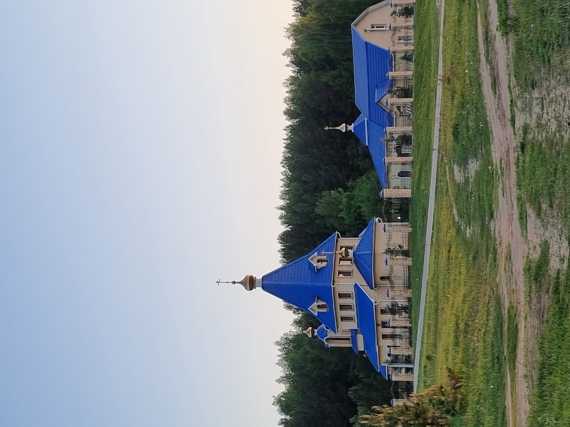 A sideways view captures a church in Chaikovsky, Russia. The church is constructed in light-colored brick and features a bright blue roof with pointed domes. Golden crosses stand atop the domes. The building is surrounded by a grassy field and a line of trees, with a gravel road running parallel to the church. The sky is a pale blue.