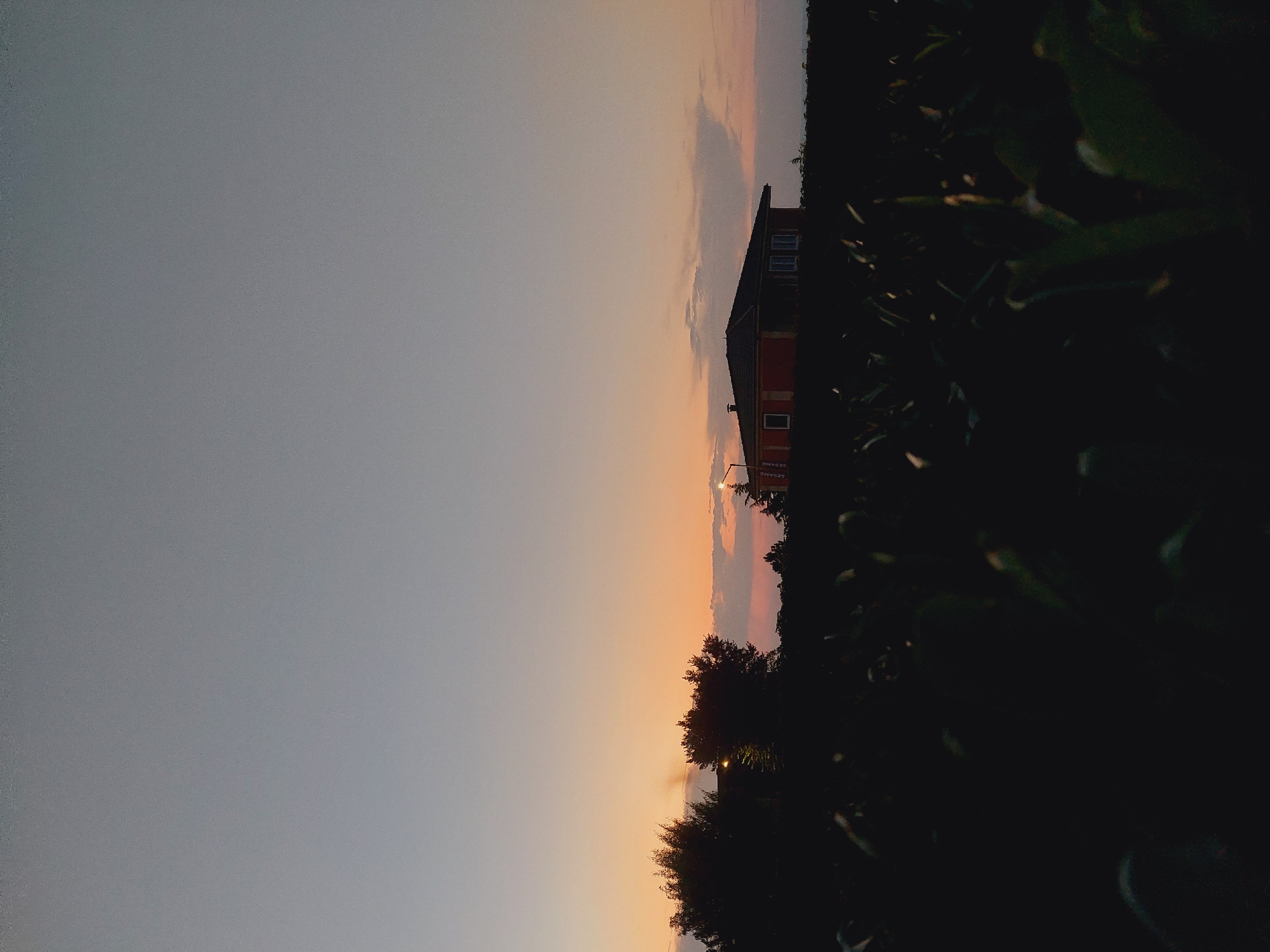 This image depicts a rural landscape at dusk. In the foreground, a dark, indistinct field of vegetation occupies the lower portion of the frame. In the mid-ground, a single-story house with a dark roof and red walls is visible. The house has several windows, some of which appear to be lit. To the left of the house, silhouetted trees stand against the twilight sky. A street lamp is present near the house. The sky above transitions from a pale blue at the top to hues of orange and pink near the horizon, indicating either sunrise or sunset. The scene appears to be set outside of an urban environment, suggesting a rural or suburban location. There are no people or animals visible in the image. The overall mood is tranquil and serene. The provided location of Sozopol, Bulgaria, aligns with this depiction of a coastal town or its surrounding countryside.