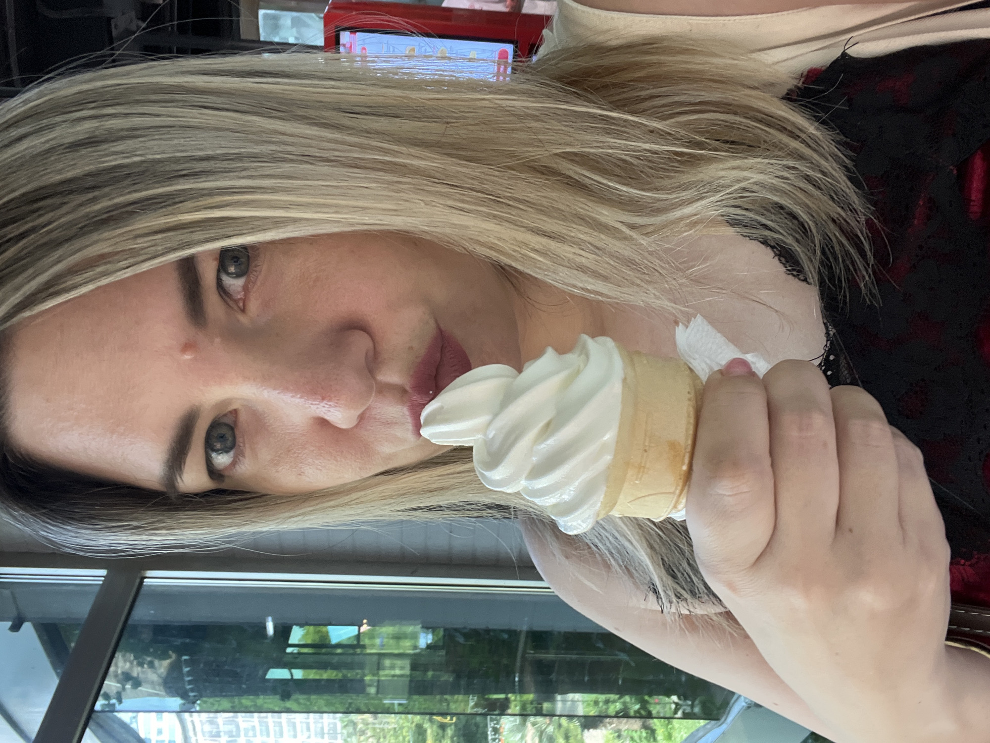 A woman with blonde hair is captured in a close-up, sideways portrait as she holds a soft-serve ice cream cone. She is wearing a black and red lace-trimmed top, and a beige strap from a bag is visible over her shoulder. The ice cream is a pale yellow and swirled in a typical soft-serve shape. Her right hand, with manicured nails, holds the cone. The background appears to be outdoors, with reflections of trees and buildings visible in a large window or glass surface behind her. A portion of a red electronic device with some visible text is also in the upper portion of the image. The lighting suggests it is daytime. The woman's expression is neutral as she looks towards the camera with one eye visible. There is no clear indication of the specific location in Sochi, Russia, beyond the general outdoor setting and the possible reflection of urban elements.