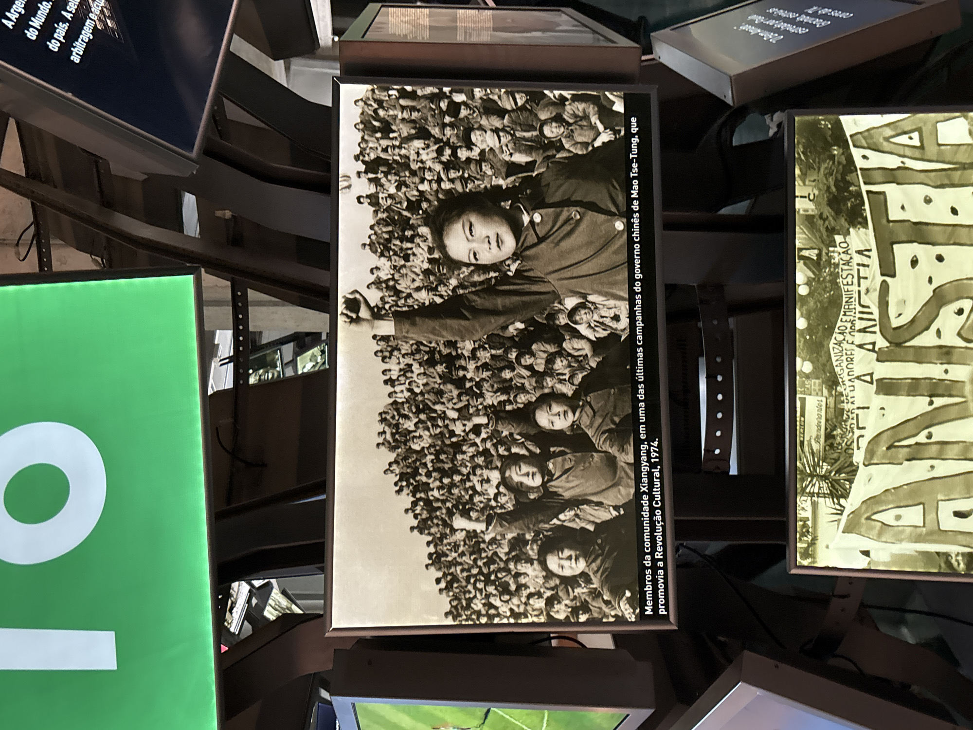 This is an image taken inside a museum, likely the Museu da Imagem e do Som (MIS) in São Paulo, Brazil, given the exhibit style. The photo depicts a display featuring a black and white photograph of a crowd of people, specifically "Membros da comunidade Xiangyang, em uma das últimas campanhas do governo chinês de Mao Tse-Tung, que promovia a Revolução Cultural, 1974". The image shows several young people with arms raised in a show of solidarity.
Several other informational displays are visible around the central photo. One display has the word "O" in large white letters on a green background. Another display contains text with the words "Mundo," "país," and "arbitragem." Another display contains the text "ORGANIZAÇÃO E MANIFESTAÇÃO ANISTIA."
The scene shows that the photograph is being exhibited as part of a cultural display in the museum.