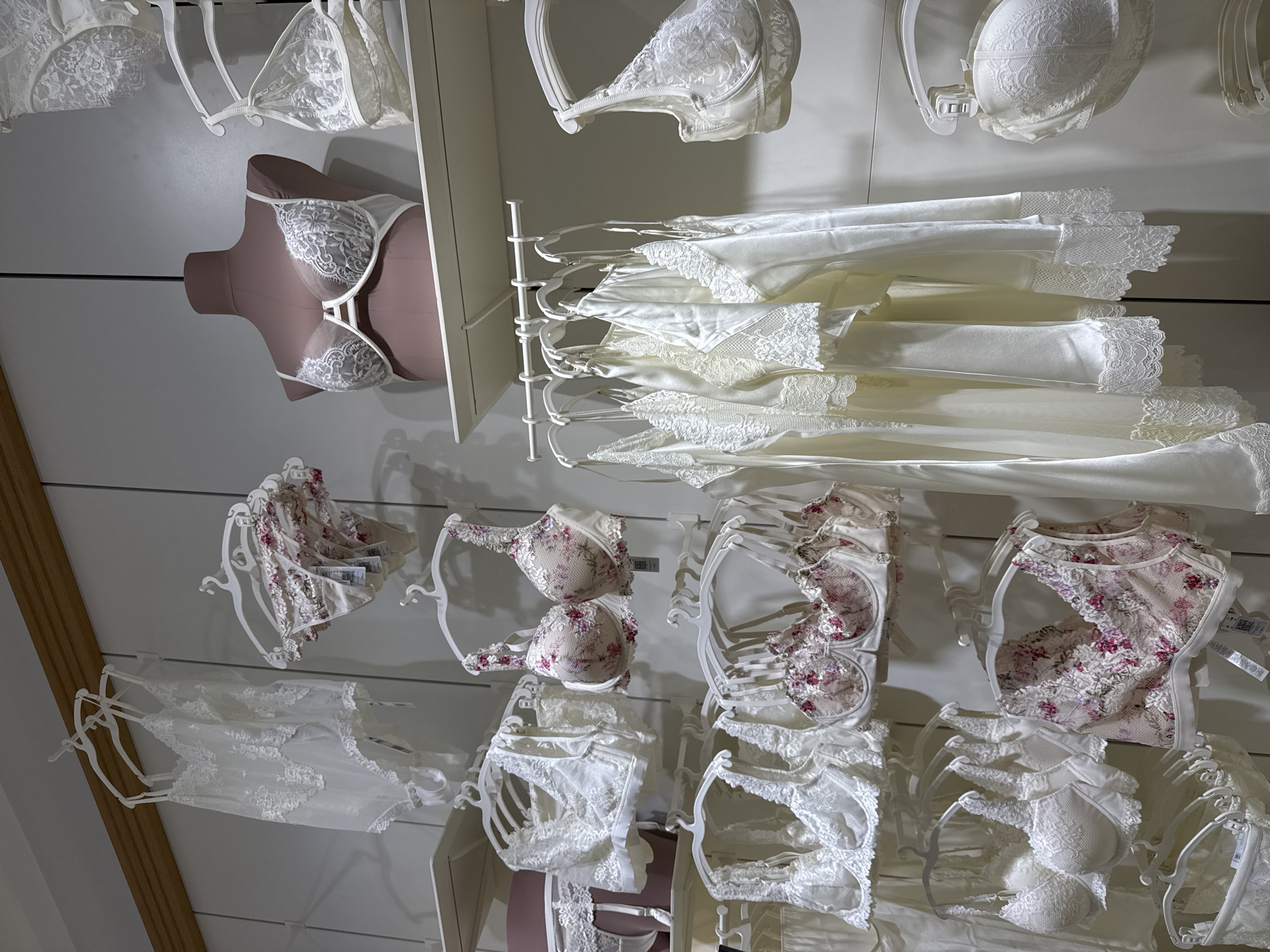 The image shows a display of women's lingerie and nightwear on a white wall, likely inside a store. The items are neatly arranged on hangers. At the top are several white lace bras and satin slips. Below, there is a pink mannequin with a white lace bra. Lower, there are floral-patterned lingerie sets along with more white lace bras and panties. The tags are visible on some of the items.