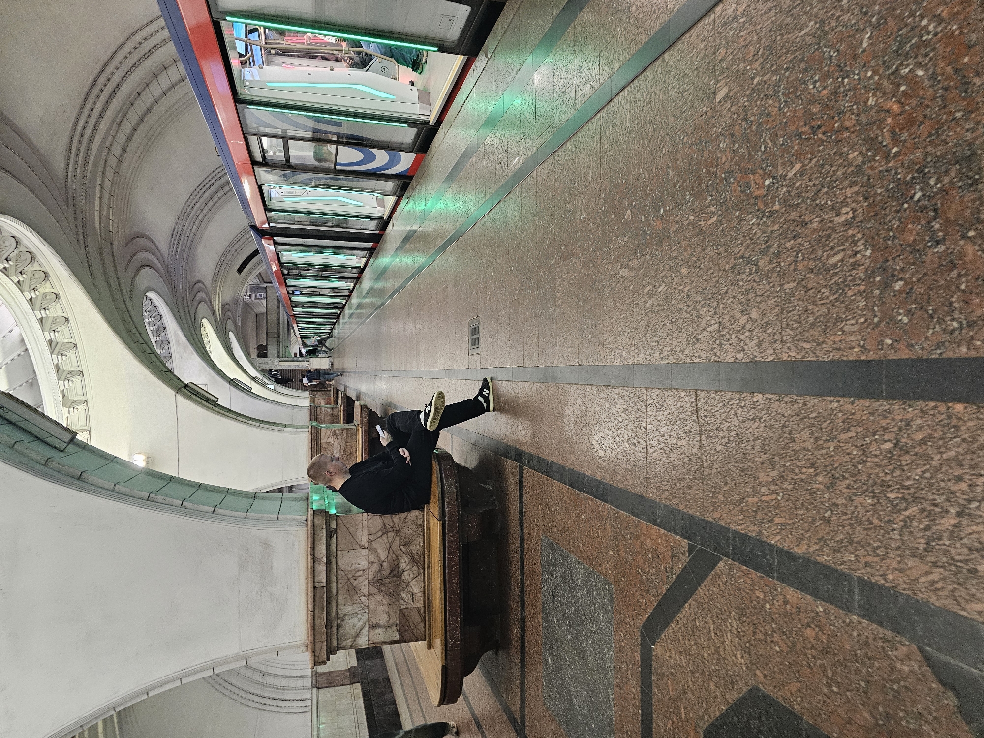 A man sits on a wooden bench in a Moscow metro station, looking at his phone. He is wearing a black sweater and black pants with white-striped sneakers. The station is ornate with white arched ceilings and marble pillars. A train with bright green lighting along its side is stopped at the platform, with its doors open. The floor is made of speckled red and black granite.