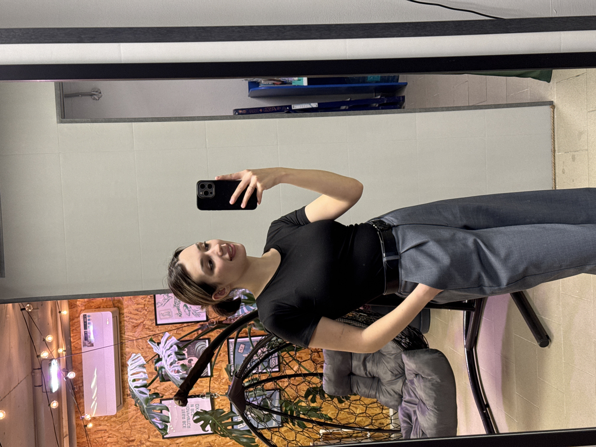 A woman is taking a selfie in a mirror. She is wearing a black top and grey trousers with a black belt. She is holding a black smartphone in her left hand. Behind her is a hanging chair with grey cushions, positioned in front of a wall decorated with artwork and string lights. An air conditioning unit is mounted on the wall. The scene appears to be indoors, possibly in a stylishly decorated room. A white tiled wall with a shelf is visible at the top of the mirror's reflection. The location is in the city of Unknown, Unknown.