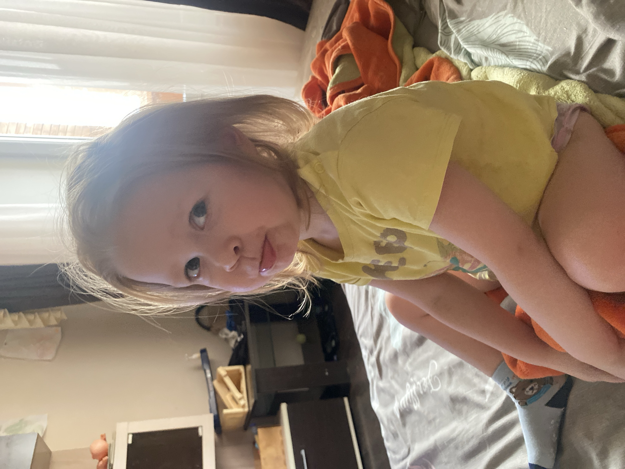 The image depicts a young child seated on a bed in an indoor setting located in Chaikovsky, Russia. The child, with light-colored hair and blue eyes, faces the camera with their tongue slightly extended from the mouth. The child wears a yellow short-sleeved t-shirt displaying dark markings, light pink undergarments, and a light blue sock with a brown bear design on the left foot. An orange blanket or towel is draped over the child's lap and extends to the side. The bed is covered with a light grey duvet featuring a subtle pattern. In the left background, a white shelving unit holds a dark-screened device and a small orange object, with a dark brown cabinet or desk below it supporting wooden boxes. To the right, a bright window is visible, covered by sheer white curtains, showing a brick building exterior under daylight conditions.