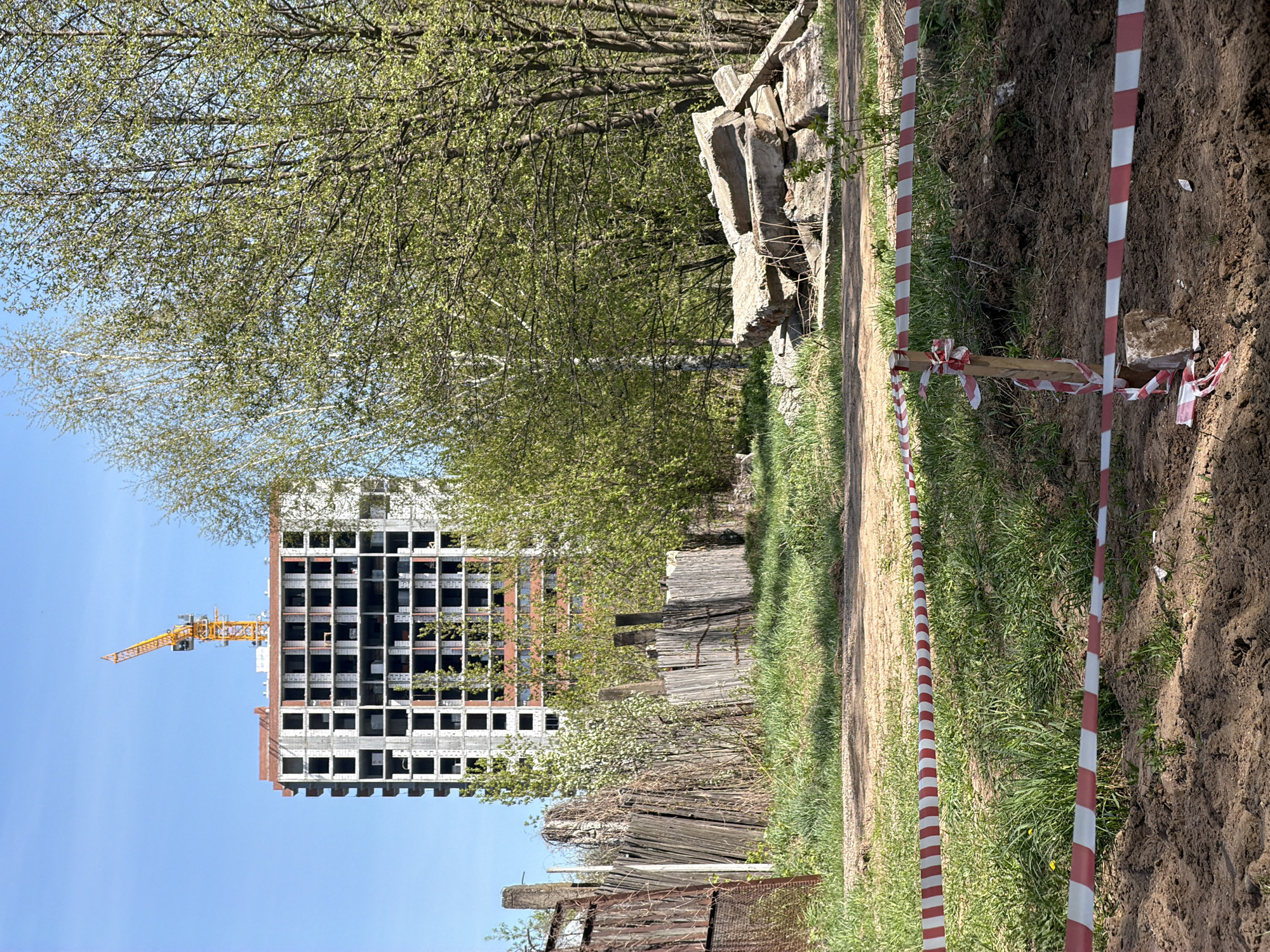 The image shows a construction site near Chaikovsky, Russia. An unfinished high-rise building with a yellow crane is visible in the background under a clear, blue sky. A dirt road runs through the foreground, bordered by overgrown grass and marked by red and white striped warning tape on stakes. An old, weathered wooden fence is on one side of the road, while a recently excavated dirt ditch is on the other side. Piles of concrete debris are on the side opposite the building. Trees are visible behind the construction site and along the edges of the road. The photograph is taken from a ground-level perspective.
