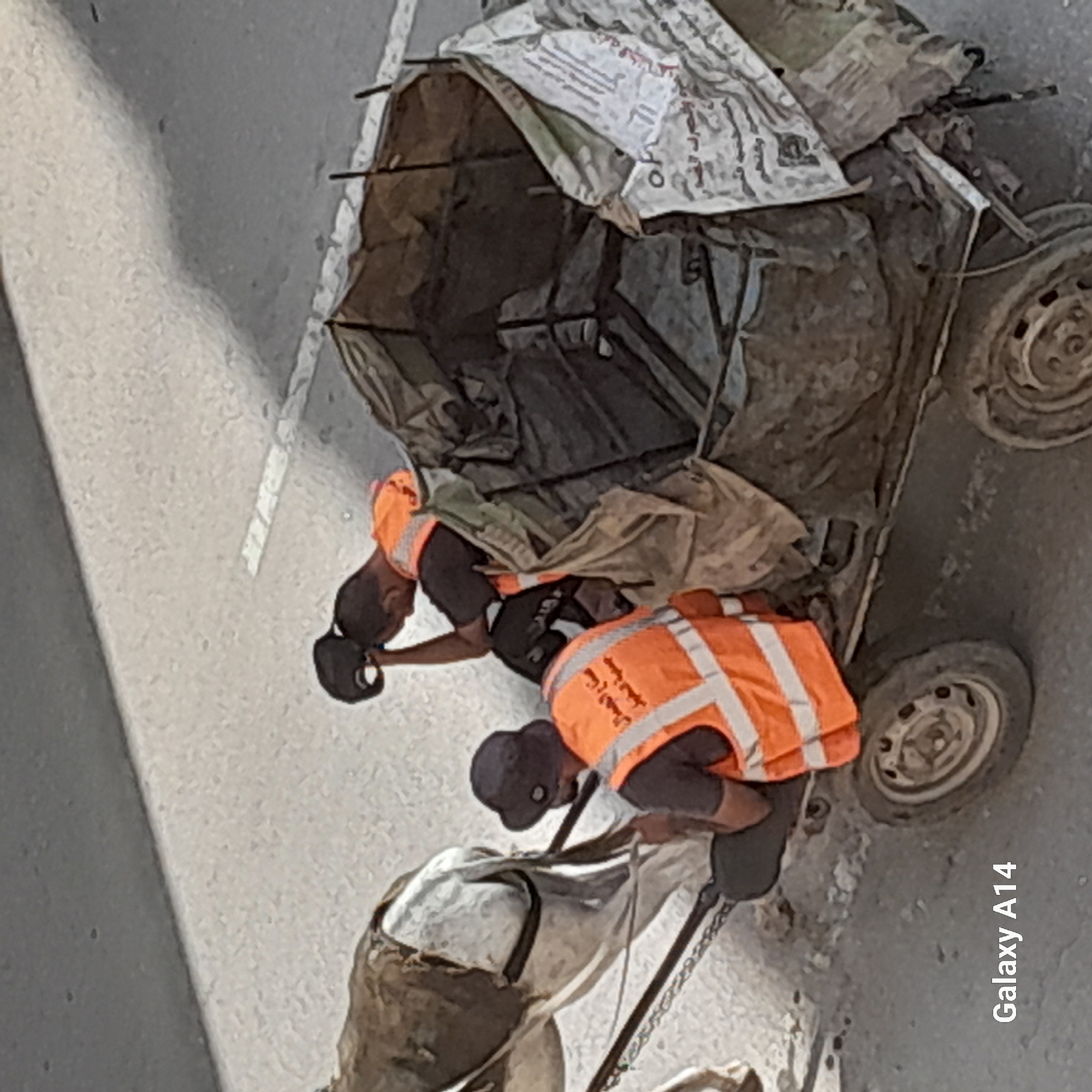 The image shows two men wearing orange reflective vests and dark hats, sitting on a horse-drawn cart filled with trash. One man is holding his head in his hand, appearing weary. The cart has a metal frame covered with a tarp and some visible debris inside. The scene is set on a paved road, with a gray sidewalk-like surface to the upper left. The bottom left corner of the image contains the text "Galaxy A14." The image appears to be taken in the city of Khan Yunis, Palestinian Territories.