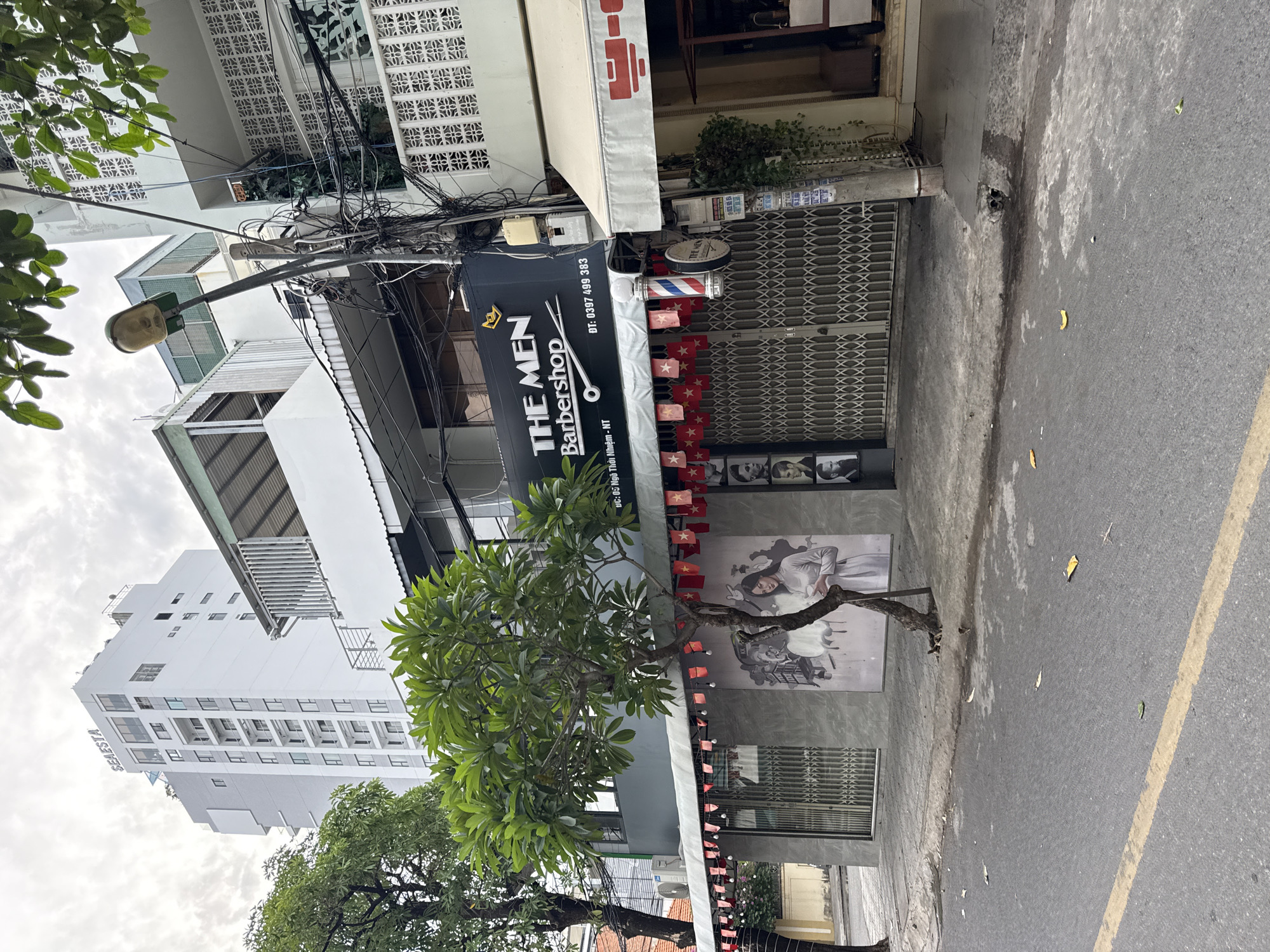 The image shows a street scene in Nha Trang, Vietnam. In the foreground, a grey asphalt road with a yellow line running down the left side. The sidewalk is visible, with a tree growing from it. Behind the sidewalk, a building with a grey facade and a large black sign that reads "THE MEN Barbershop" is prominent. Below the sign, a mural depicts a woman in traditional Vietnamese attire. The building has a metal grate door and is decorated with red and white flags. To the right of the barbershop, a building with a light-colored awning is partially visible. In the background, a tall white building with the word "SESTA" on top is seen. Overhead, a cloudy sky is visible, with some trees and electrical wires adding to the urban landscape.
