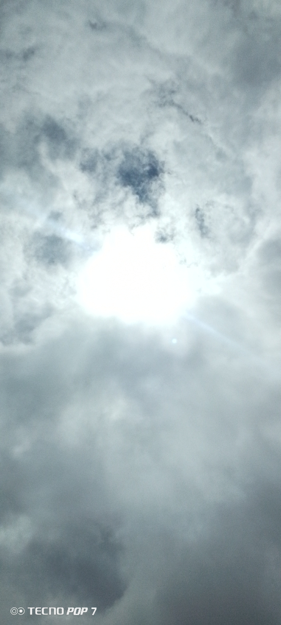 A photograph taken from below shows a bright sun shining through a cloudy sky. The sun is a white, blown-out disc in the center of the frame, with rays of light emanating outwards. The clouds are a mix of light and dark grays, with some areas appearing wispy and others dense. The bottom right corner of the image contains a watermark that reads "TECNO POP 7". The image is undated and unlocated, but the prompt suggests the location is Altagracia, Venezuela.