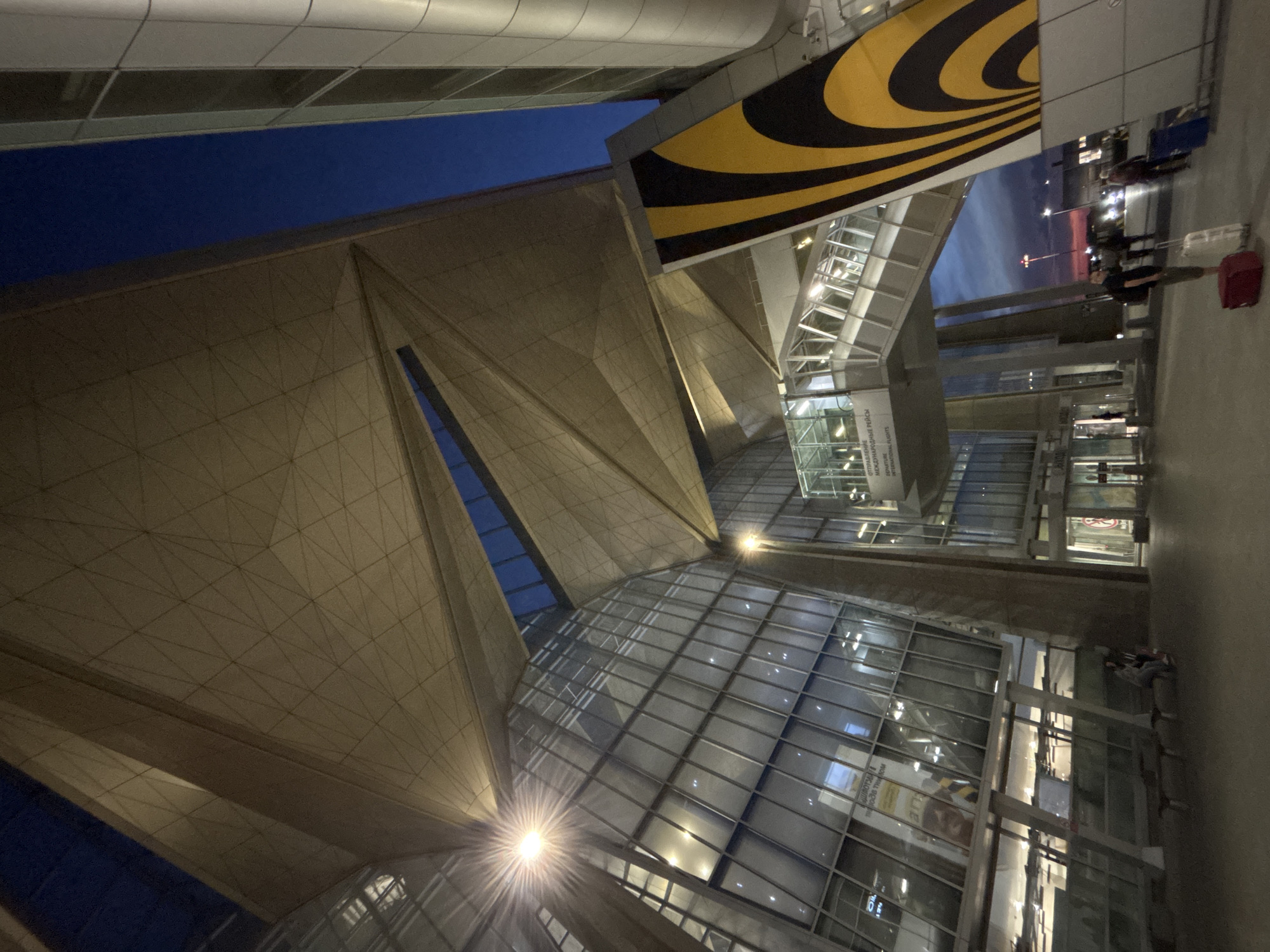 The image depicts the exterior of Pulkovo Airport in Saint Petersburg, Russia, at dusk or twilight. The scene is dominated by the modern, angular architecture of the terminal building, characterized by a large, light-tan geometric canopy featuring multiple facets and extensive glass curtain walls. The sky transitions from a deep blue overhead to an orange and pink hue at the horizon on the right.
Artificial lighting is active, with bright interior lights visible through the glass facades and exterior streetlights, one of which on the left exhibits a prominent starburst effect. A pedestrian skybridge connects parts of the terminal, identifiable by its distinctive black and yellow striped paneling on the right side. A white sign mounted on the skybridge is visible, bearing the text "ОТПРАВЛЕНИЕ МЕЖДУНАРОДНЫЕ РЕЙСЫ" and "DEPARTURE INTERNATIONAL FLIGHTS."
On the paved ground level in the foreground, several individuals are present. To the far left, two people are seated on a dark bench. In the lower right corner, a person wearing a dark top and backpack stands beside a red carry-on suitcase and a white upright suitcase. Behind them, another individual walks away from the viewer, pulling a blue upright suitcase. Additional faint figures and vehicle lights are visible in the distance towards the horizon.