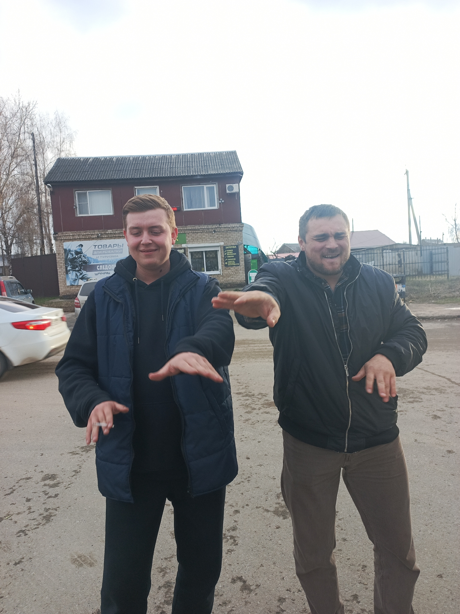 Two men are captured in an outdoor setting, likely a street or parking area, in front of a two-story commercial building. The man on the left, with short light brown hair, wears a black hoodie under a dark blue quilted vest and black pants. He smiles broadly with his eyes partially closed and holds a lit cigarette between the fingers of his left hand. Both his hands are extended forward, palms down, in a gestural pose.
To his right, the second man, with a beard and darker hair, wears a dark zippered jacket over a plaid shirt and brown trousers. He also smiles and laughs, with his hands extended forward in a similar pose as the first man. Both individuals appear to be in good spirits, possibly engaged in a shared joke or activity.
The background features the commercial building, which has multiple windows and a gray-tiled roof, with an air conditioner unit visible on its right side. A large sign on the building displays Cyrillic text: "ТОВАРЫ ДЛЯ РЫБАЛКИ И ТУРИЗМА" (Goods for Fishing and Tourism) with "ЯМАН" (YAMAN) below it, indicating a shop. Another sign on the right, partially visible, reads "ЗАТОЧКА" (Sharpening) followed by "НОЖЕЙ" (Knives), "ОХОТНИЧЬИХ" (Hunting), "ТОПОРОВ" (Axes), and "БЕНЗОПИЛ" (Chainsaws), suggesting a sharpening service. A digital clock above this sign shows "09", possibly indicating time.
Behind the men, a white car is partially visible on the left, and various trees, bare of leaves, along with utility poles, frame the scene under a bright, overcast sky. The ground is a mix of asphalt and dirt. The general setting suggests a daytime scene in a small town or city, consistent with the provided location context of Chaikovsky, Russia.