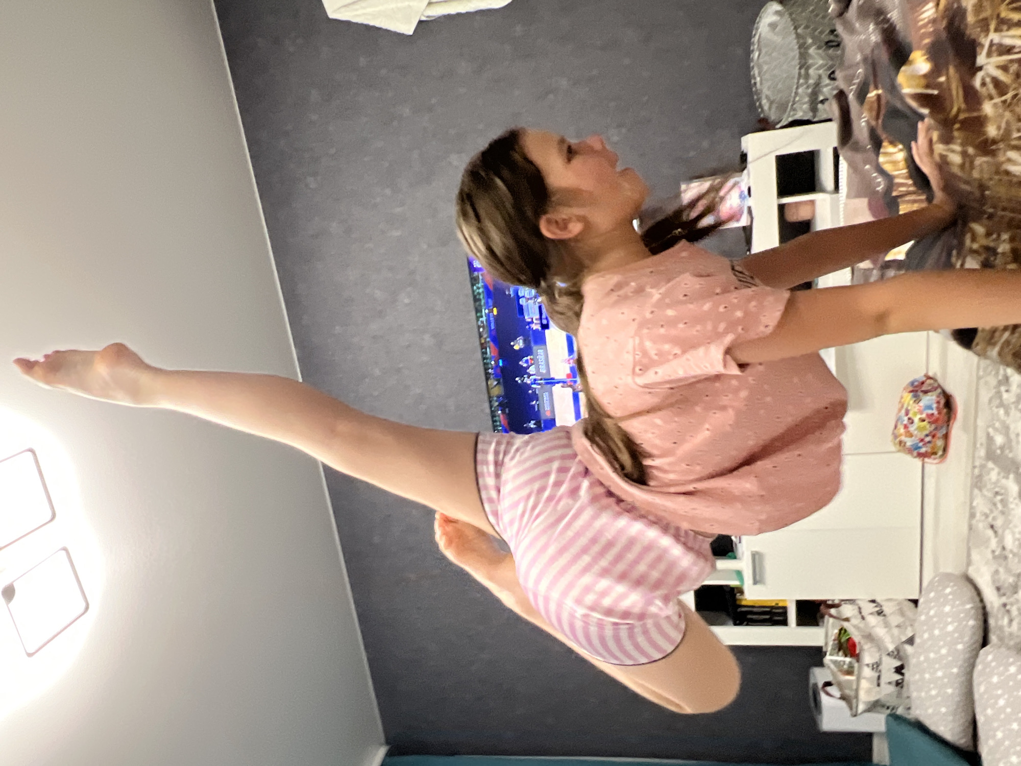 A young female performs a gymnastic pose in an indoor residential setting in Chaikovsky, Russia. Her right leg is extended vertically towards the white ceiling, which features two illuminated rectangular light fixtures. She is wearing pink-and-white striped shorts and a light pink short-sleeved top with a subtle pattern. Her body is bent at the waist, and her left arm is planted on a bed or sofa covered with a brown and gold patterned blanket, supporting her weight. Her head is turned towards the right, showing her profile. In the background, a large flat-screen television displays what appears to be a sports event with a "BRASDAR" banner visible. The room features a dark grey textured wall, a white wall-mounted cabinet system with various items, a light-colored rug on the floor, and several decorative pillows. A grey patterned laundry basket and a colorful fabric item are also visible on the floor.