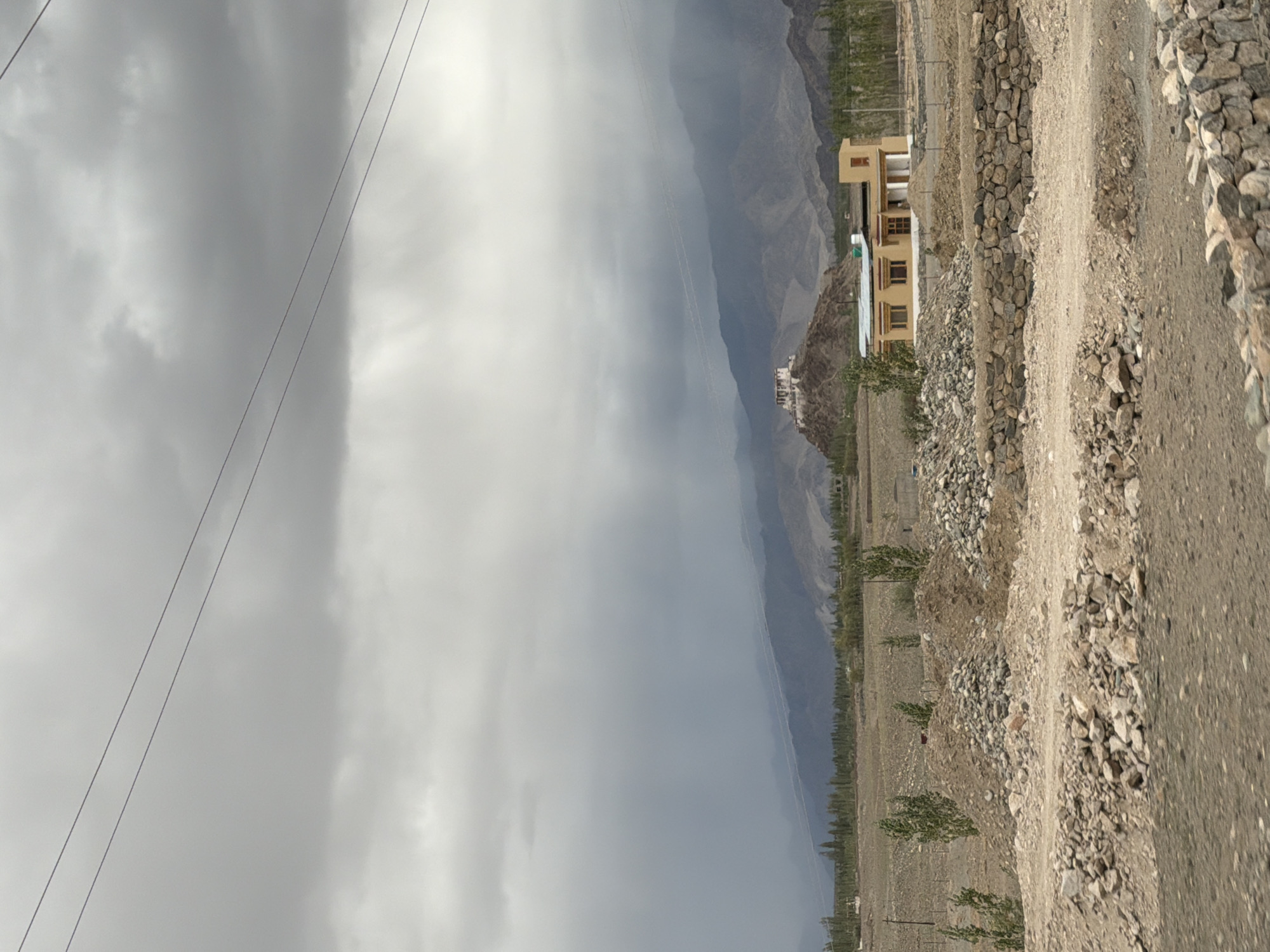The image depicts a panoramic view of a rural landscape under a heavily overcast sky. In the distance, a cluster of buildings, possibly a monastery, is perched atop a rocky hill. The surrounding terrain is mountainous and arid, with scattered vegetation. In the foreground, there are piles of rocks and a stone wall, suggesting construction or land clearing. A single-story building with a yellow facade and a grey roof is also visible in the mid-ground. The scene is devoid of any people or animals, creating a sense of stillness and isolation. The location is identified as Tanglakpurna, India.