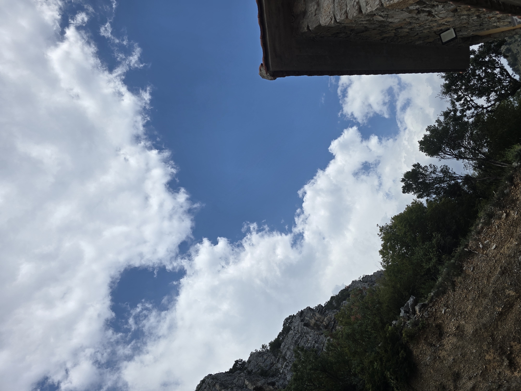The image captures a close-up view of the sky with scattered clouds and a rocky mountainside on the left. A portion of a stone building with a tiled roof is visible on the right side of the frame. The building appears to have a modern outdoor light fixture attached to its wall. The environment suggests a natural setting, possibly a rural or mountainous area, with vegetation growing on the slope. There are no people or animals depicted in the image. The lighting indicates it is daytime. Without further information or visual cues, it is not possible to definitively place this scene within the city of Podgorica, Montenegro, though it could be a depiction of a structure or landscape found in its surrounding region.