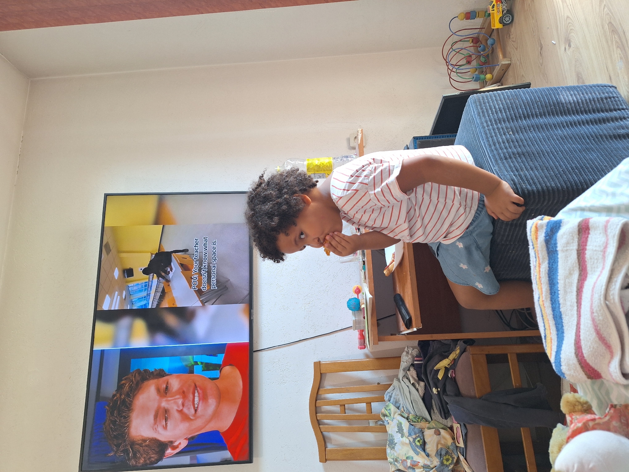 In a dimly lit room in Blagoevgrad, Bulgaria, a young child with curly hair sits on a blue ottoman, eating a piece of food. The child is wearing a red and white striped t-shirt and blue shorts. To the left, a television screen displays a video with a man's face on the left side and a classroom scene on the right, with text that reads "POV: Your teacher doesn't know what personal space is." A wooden chair filled with clothes is next to the television. A small wooden desk with various items, including a remote control, a bottle of water, and some toys, is positioned in front of the child. In the background, a bead maze toy and a small yellow toy truck are visible on the wooden floor. The room appears to be a living space, possibly a bedroom or play area.