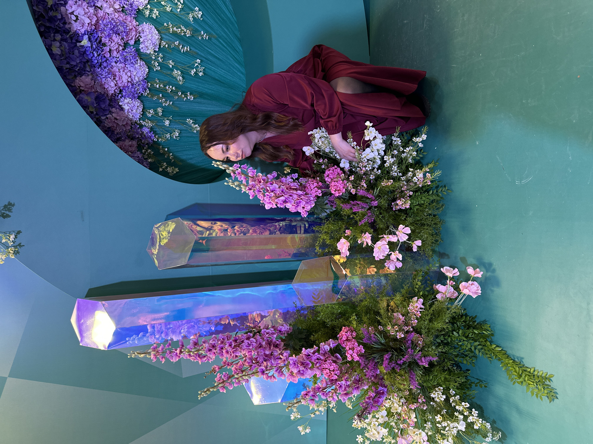 A photo of a woman in a red dress lying on a green surface, surrounded by purple and white flowers. The woman is leaning on her left side, with her head resting near the flowers. Her right hand rests near the flowers. There are large hexagonal-shaped structures partially covered in flowers in front of the woman. The background is green. The location is unknown.