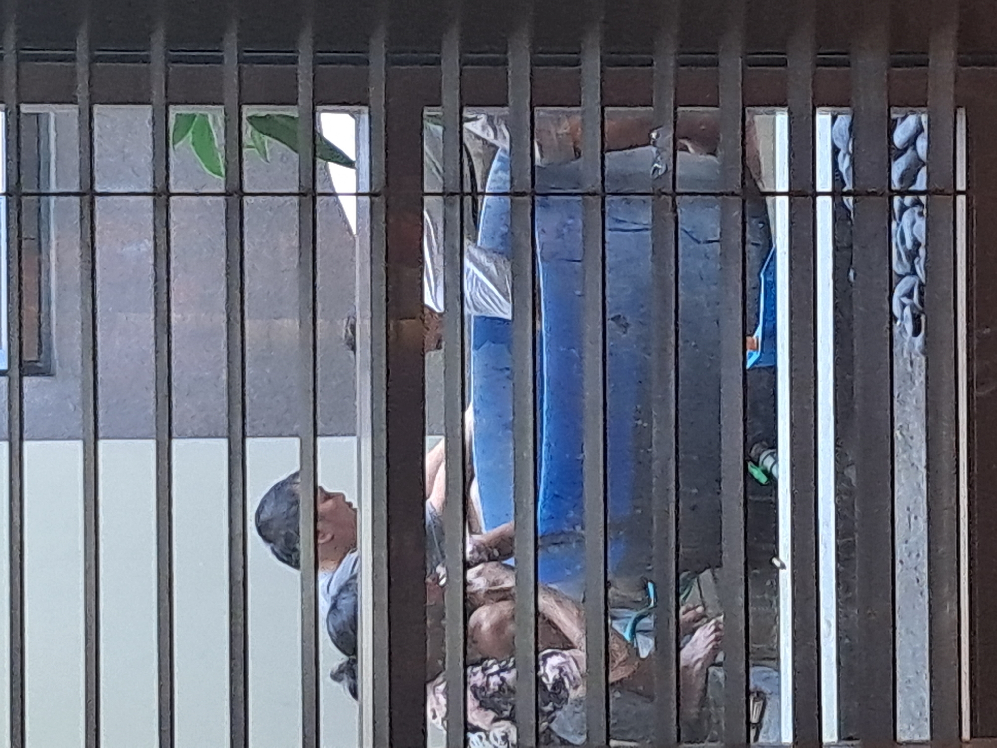 The image, taken through blinds, depicts an outdoor scene in Bareng Lor, Indonesia, where several people are gathered around a large blue tub. One individual, appearing to be a young man with dark hair tied back, is visible from the shoulders up, looking down and potentially interacting with the tub's contents. Another person, partially obscured, is also near the tub. The tub itself appears to be filled with water, and there are reflections of people and patterns on its surface, suggesting it is large and possibly used for bathing or a similar activity. To the left of the tub, a patterned cloth or fabric is visible, draped over something or someone. In the foreground, a blue toy boat rests on the ground near the tub. The background shows a textured wall and some greenery, with sunlight filtering through the blinds, creating horizontal bands of light and shadow across the scene. The ground around the tub is covered with small, rounded stones.