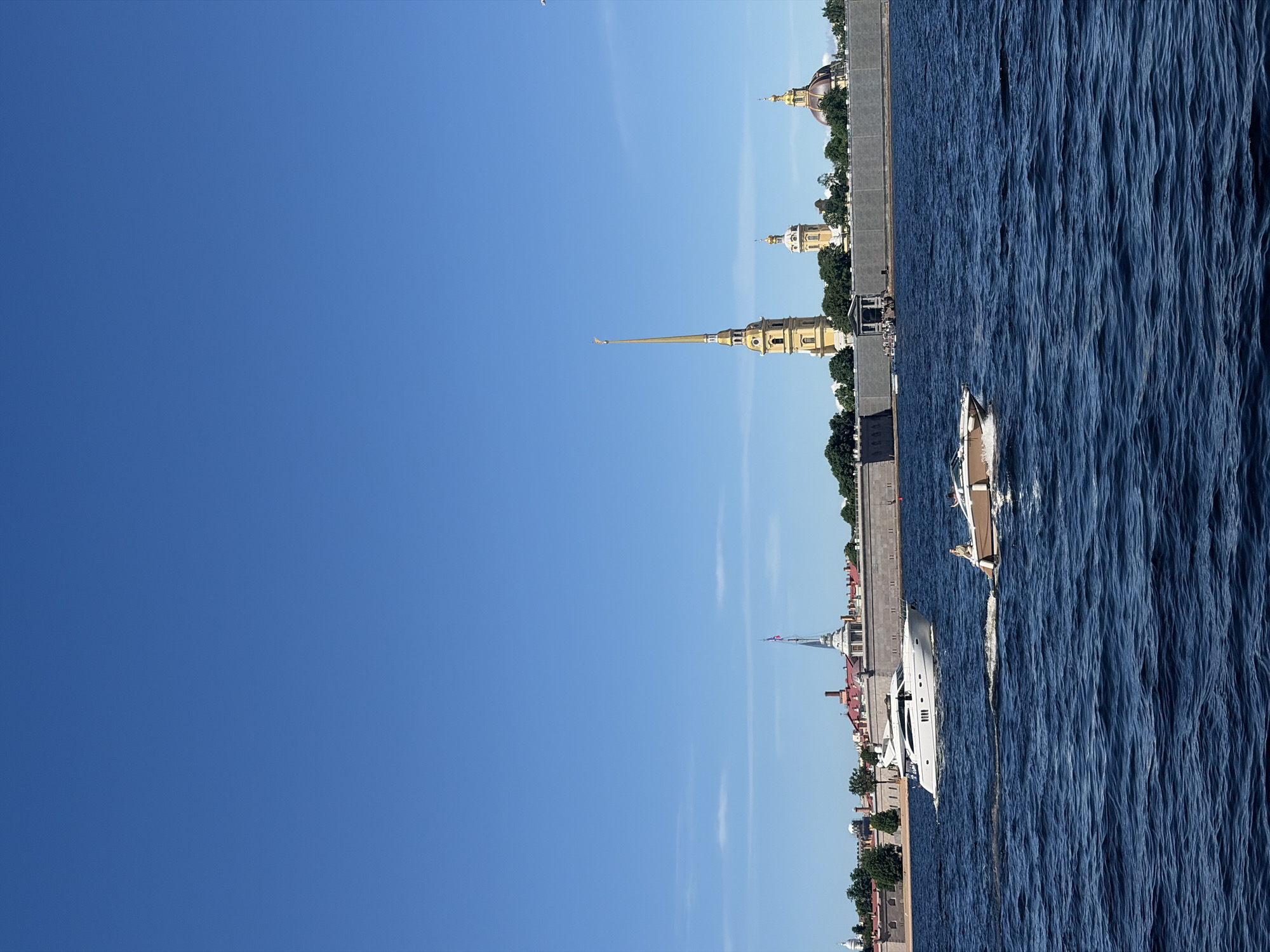 The image depicts a daytime scene on the Neva River in Saint Petersburg, Russia, with the Peter and Paul Fortress in the background under a clear blue sky.
The foreground is dominated by dark blue, choppy water with visible ripples and small waves. Two motorboats are in motion. A large, white motor yacht with multiple dark windows is positioned slightly left of center, moving from left to right, leaving a discernible wake. Closer to the viewer and to the right, a smaller, light brown and white motorboat is also moving, creating a wake. One person, wearing light-colored attire, is visible seated at the bow of the smaller boat, facing toward the right side of the frame.
The mid-ground features a continuous, grey stone embankment or fortress wall running horizontally across the river, separating the water from the land. In the central section of this wall, a stone archway or gateway leads up from the water, with a group of people visible on steps or a platform within the opening.
The background is dominated by the structures of the Peter and Paul Fortress. From left to right: a white spire with a red, white, and blue flag (likely the Russian flag) is visible behind the wall. Further to the right, the distinct tall, slender, yellow spire of the Peter and Paul Cathedral rises prominently, topped with a golden element. To its right, two buildings with golden domes are visible, set amidst green trees. The roofs of various fortress buildings are visible in shades of red-brown, with light-colored walls. The sky above is a bright, clear blue with a few faint, wispy white clouds present in the upper left portion of the frame.