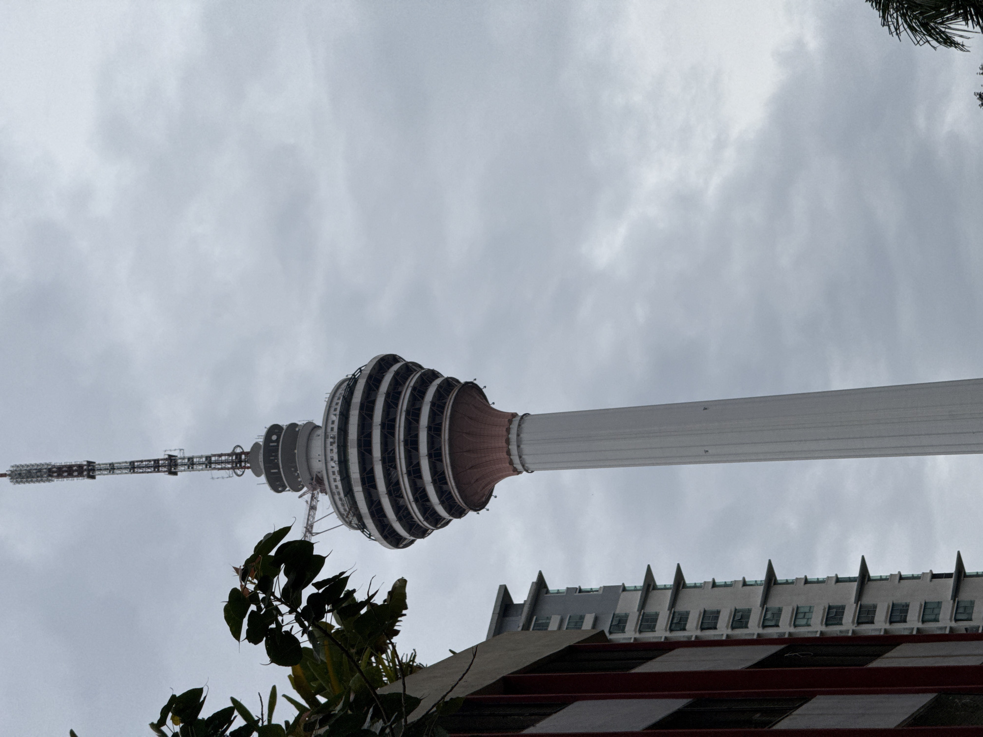 The image presents a low-angle view of the Kuala Lumpur Tower (Menara Kuala Lumpur) against a cloudy sky. The tower's distinctive bulbous structure, divided into multiple horizontal segments, is prominently featured. A white shaft extends from the bulbous top, and a thinner antenna rises above it. In the foreground, foliage from a tree partially obscures the view. Beneath the tower, the roof and upper facade of a building are visible, featuring a repeating pattern of windows and pointed architectural elements. The overcast sky provides a muted background, indicating possible weather conditions. The tower stands as a central landmark, while the surrounding vegetation and buildings add context to its urban setting in Kuala Lumpur.