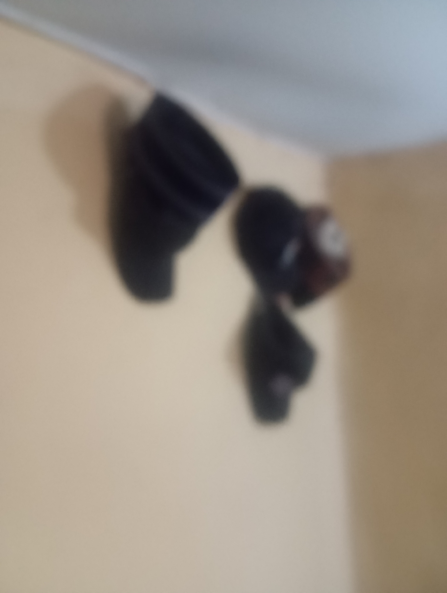The image shows a close-up, out-of-focus view of what appears to be a dark-colored fan mounted on a pale-colored wall. The fan has multiple blades that are not clearly defined due to the blurriness of the image. There is also a smaller, darker object attached to or near the fan, which is also indistinct. The lighting suggests an indoor environment with overhead illumination, possibly from a ceiling. The wall is a neutral, light beige color. The upper portion of the image shows a blurred area that could be a ceiling or the top of a doorway. There are no people, animals, or other visible objects in the scene. The image does not provide any information about the city of Jos, Nigeria, or any context that would place the scene there.