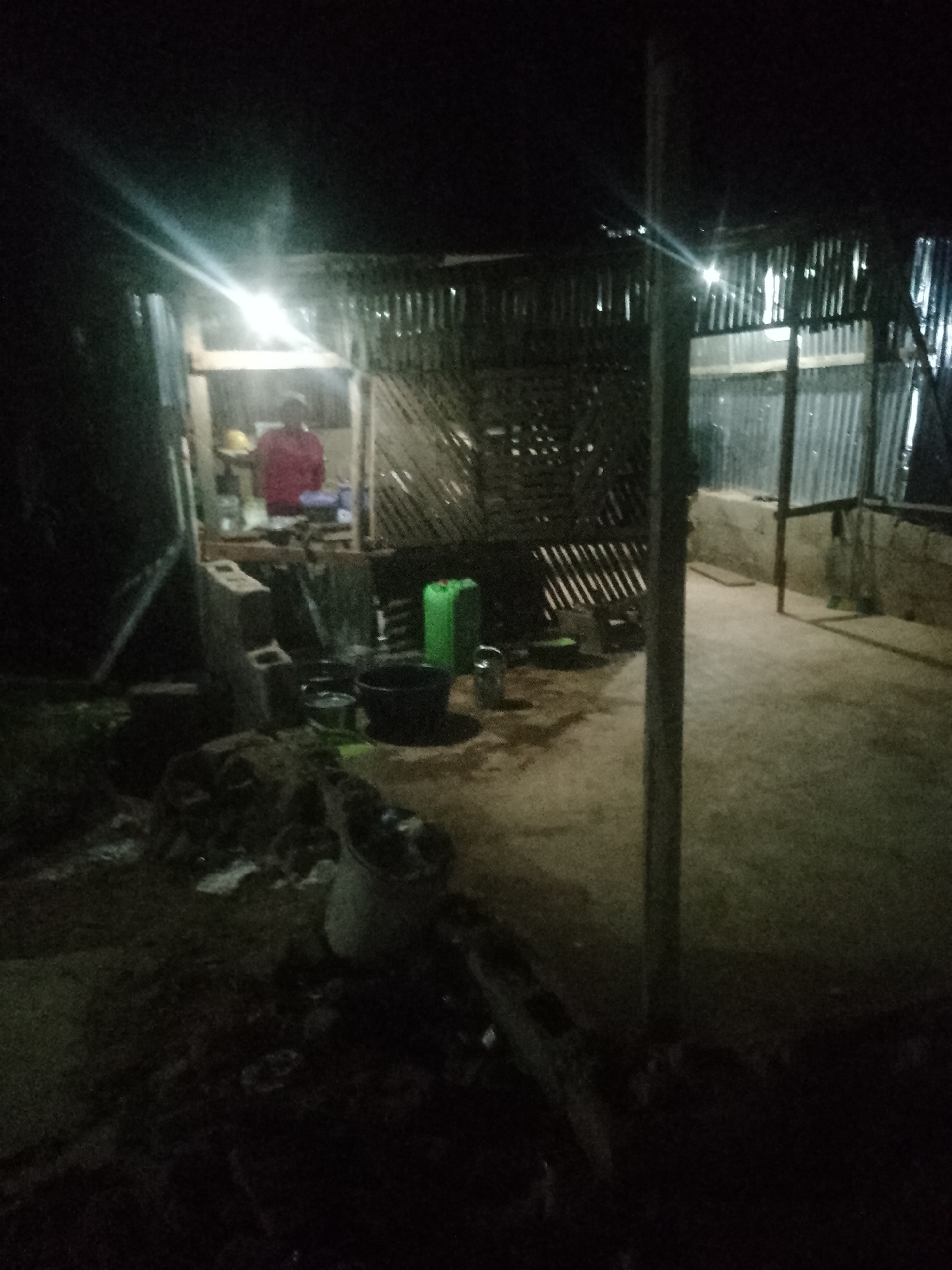 The image shows an outdoor scene at night, possibly in a rural or semi-urban setting. A person wearing a red shirt is visible inside a structure made of corrugated metal. There are various objects scattered around, including plastic containers, buckets, and what appears to be building materials. The area is dimly lit, with a bright light source creating strong glare. The ground is uneven and appears to be made of dirt or gravel. The overall impression is of a functional, but unrefined, outdoor living or working space.