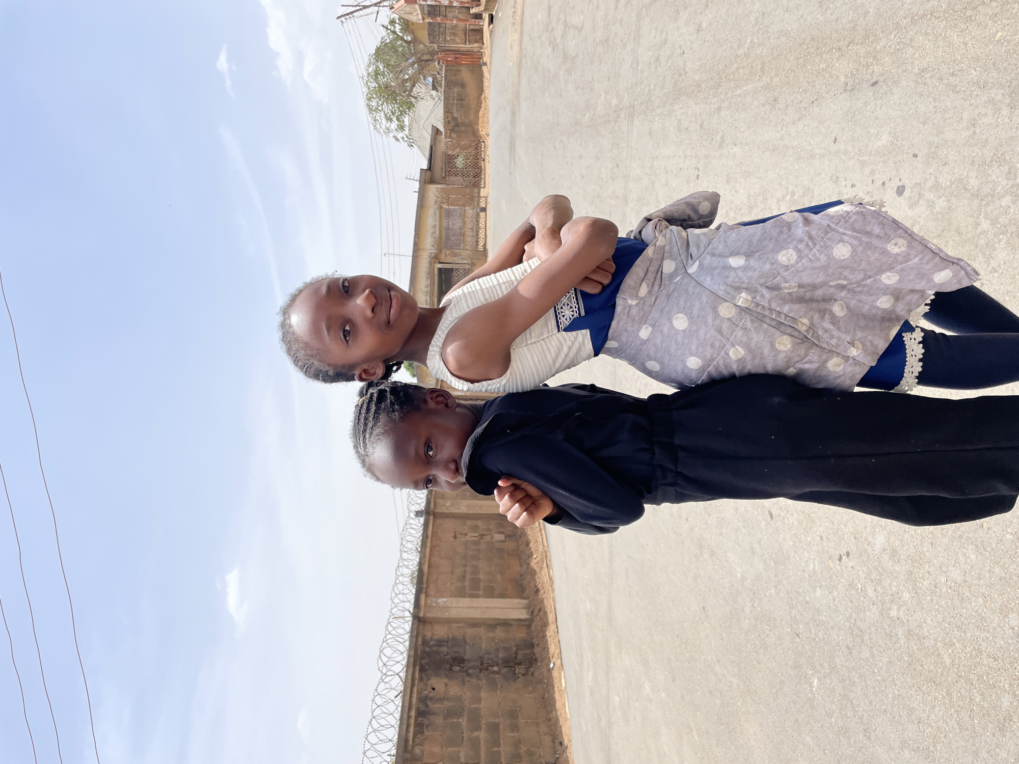 Two young girls stand back-to-back in the center of a light grey paved road under a bright, partly cloudy sky. The girl on the left, with braided hair, wears a black long-sleeved jumpsuit, clasping her hands at chest level and looking to her right (viewer's left). The girl on the right, also with braided hair, wears a white sleeveless striped top, a blue skirt, blue leggings, and a grey polka-dotted cloth tied around her waist; she stands with crossed arms, facing the camera. To the left in the background, a tan concrete block wall topped with coiled razor wire extends along the roadside. To the right in the background, multiple buildings, including what appear to be residential structures, and a sparse-leafed tree are visible. Overhead, utility lines traverse the frame. The specific location within Jos, Nigeria, cannot be determined from the visual information presented.