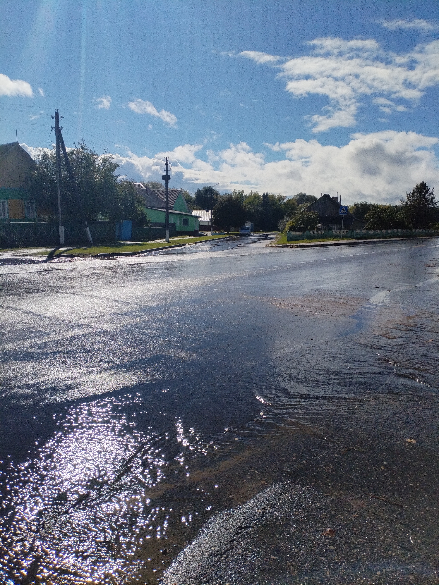 The image depicts a street scene in Bialynichy, Belarus, following a rain event. The asphalt road is wet and reflects the sky and surrounding environment. Sunlight glints off the water, creating bright, star-like patterns on the surface. On the left side of the road, houses with green and yellow facades are visible, along with a wooden fence and trees. Power lines stretch across the upper part of the frame, supported by wooden poles. In the background, more houses and trees line the street, and a pedestrian crossing sign is visible near a building. The sky is blue with scattered white clouds, suggesting that the rain has recently stopped and the sun has emerged. The overall impression is of a quiet, rural residential area experiencing the aftermath of a downpour.