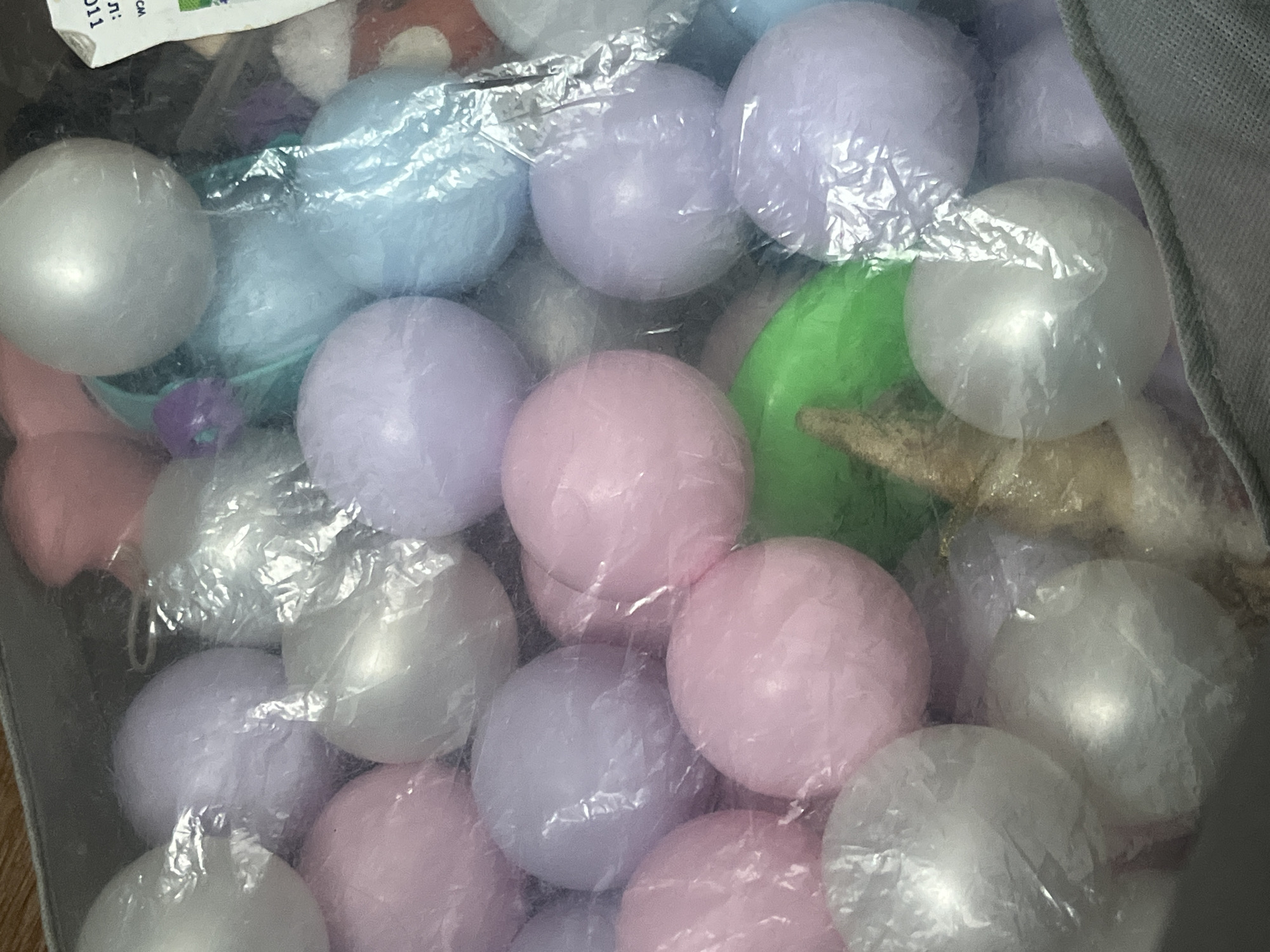The image shows a close-up, overhead view of a soft-sided storage bin filled with pastel-colored plastic balls. The balls are various shades of pink, lavender, white, and light blue, and many are partially wrapped in clear plastic packaging. A bright green ball is visible in the center of the image, and a fuzzy, cone-shaped object is partially obscured behind it. The bin appears to be made of a grey fabric material. There are no people or any identifying features of a specific city like Chelyabinsk, Russia visible in the image.