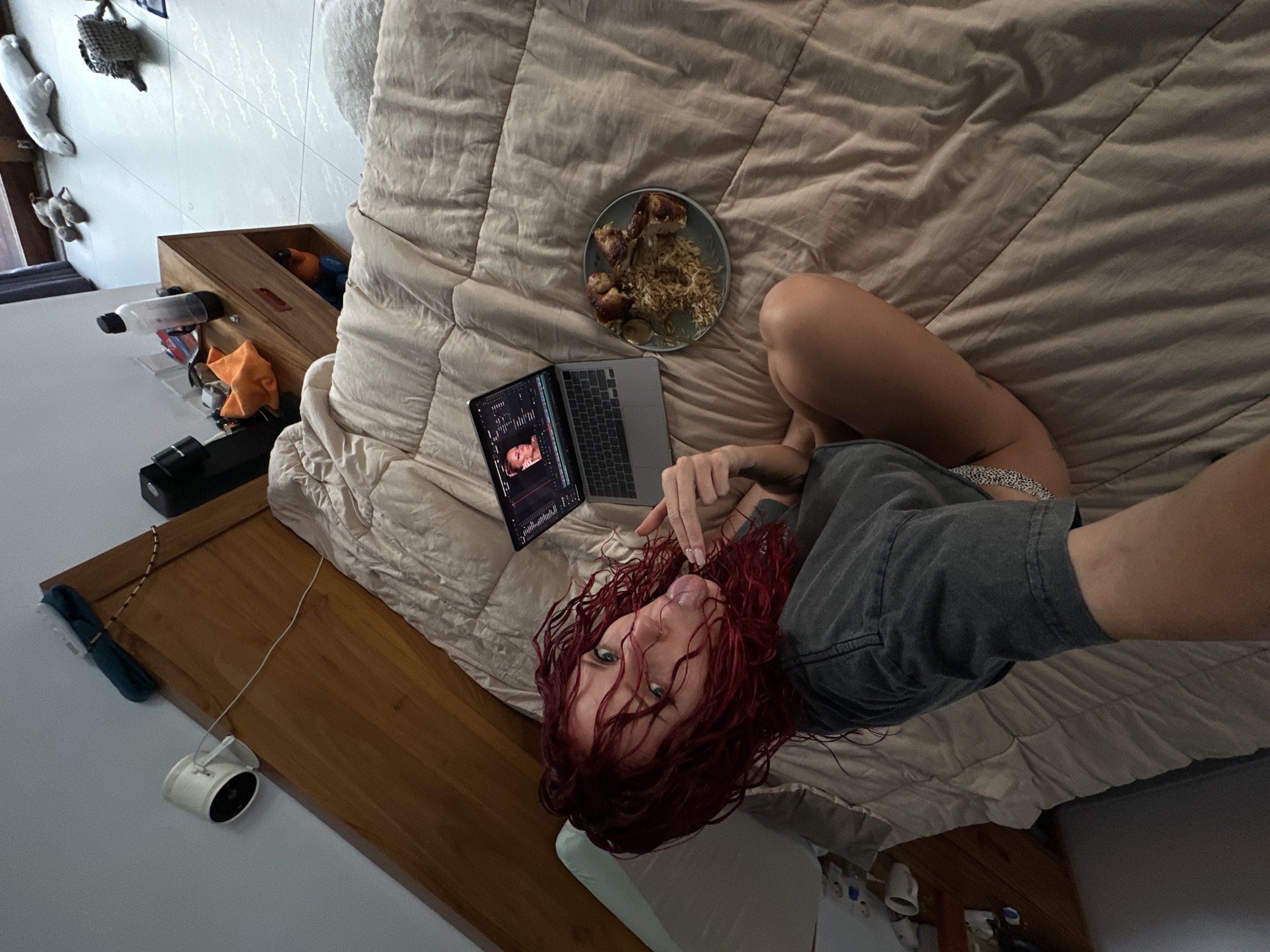 The image shows a woman with red curly hair lying on a bed in a room. She has a laptop on the bed beside her and a plate of food. A wooden bedside table is visible. The location is potentially North Kuta, Indonesia.