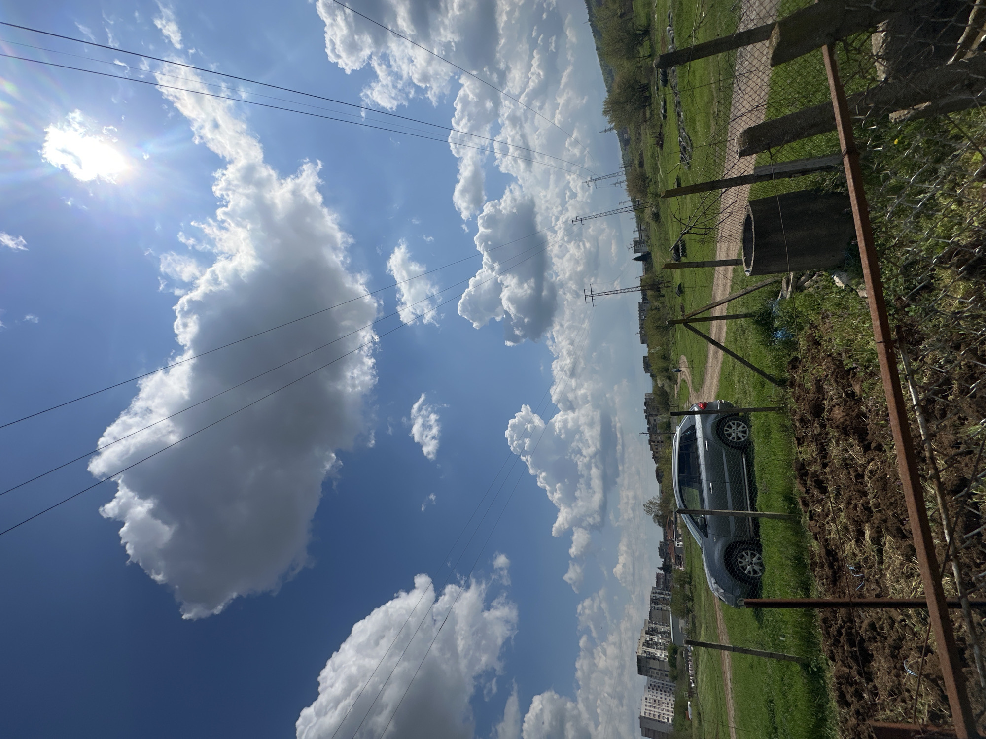 The image shows a rural scene in Montana, Bulgaria, under a partly cloudy sky with visible power lines. A silver SUV is parked beside a dirt path, near a fence and a concrete structure resembling a well. In the background, buildings indicate the edge of an urban area. The foreground features disturbed soil and overgrown vegetation.