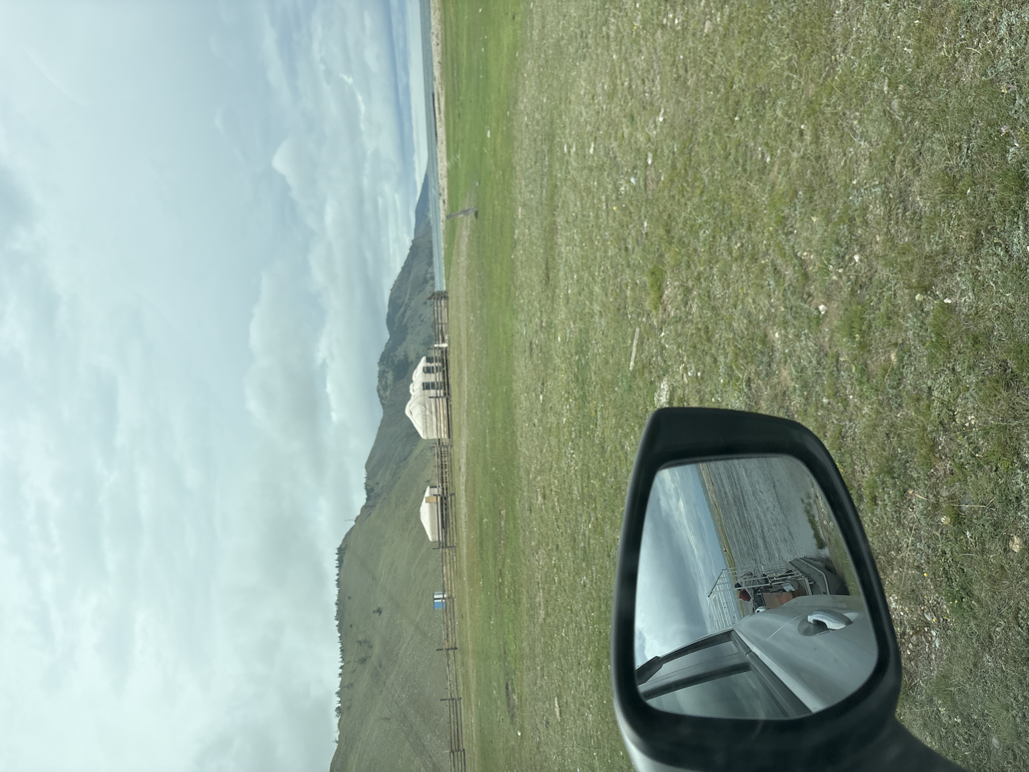 The image shows a rural landscape, likely near Bugul'deika, Russia, featuring a row of traditional yurts situated on a grassy plain. A wooden fence separates the yurts from a body of water visible in the distance. Hills covered in sparse vegetation form the backdrop under a cloudy sky. A car's side mirror in the foreground reflects a boat on the water and part of the car itself. The scene is serene and evokes a sense of natural beauty and remoteness.