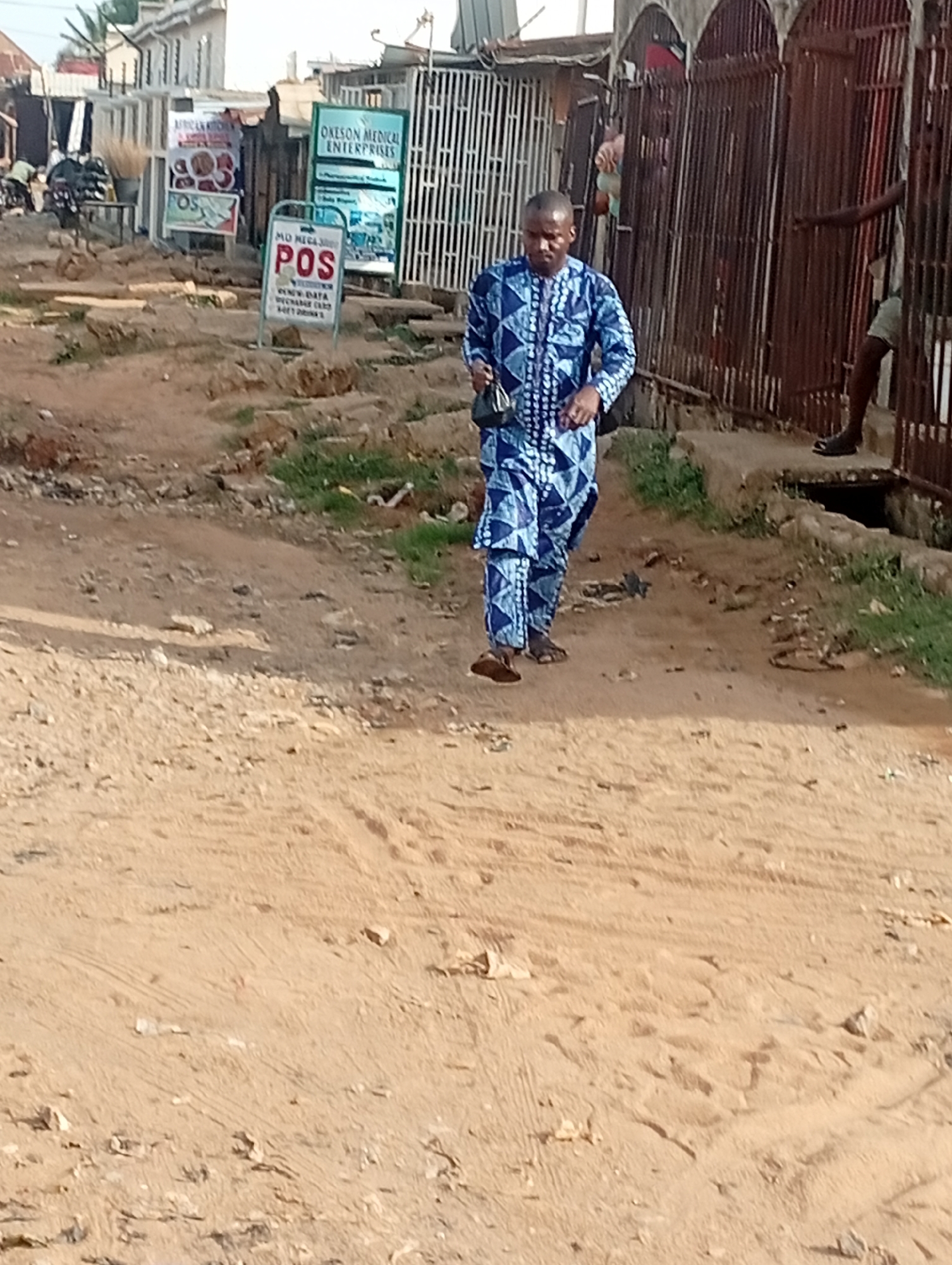 The image depicts a street in Sabon Tasha, Nigeria. A man wearing a blue patterned traditional outfit walks along the dirt road. Signs for "African Kitchen," "OKESON MEDICAL ENTERPRISES," and "POS" (Point of Sale) are visible in the background, indicating local businesses. The road appears unpaved and uneven. Buildings and fences line the sides of the road. Another man is partially visible standing near a rusted fence.