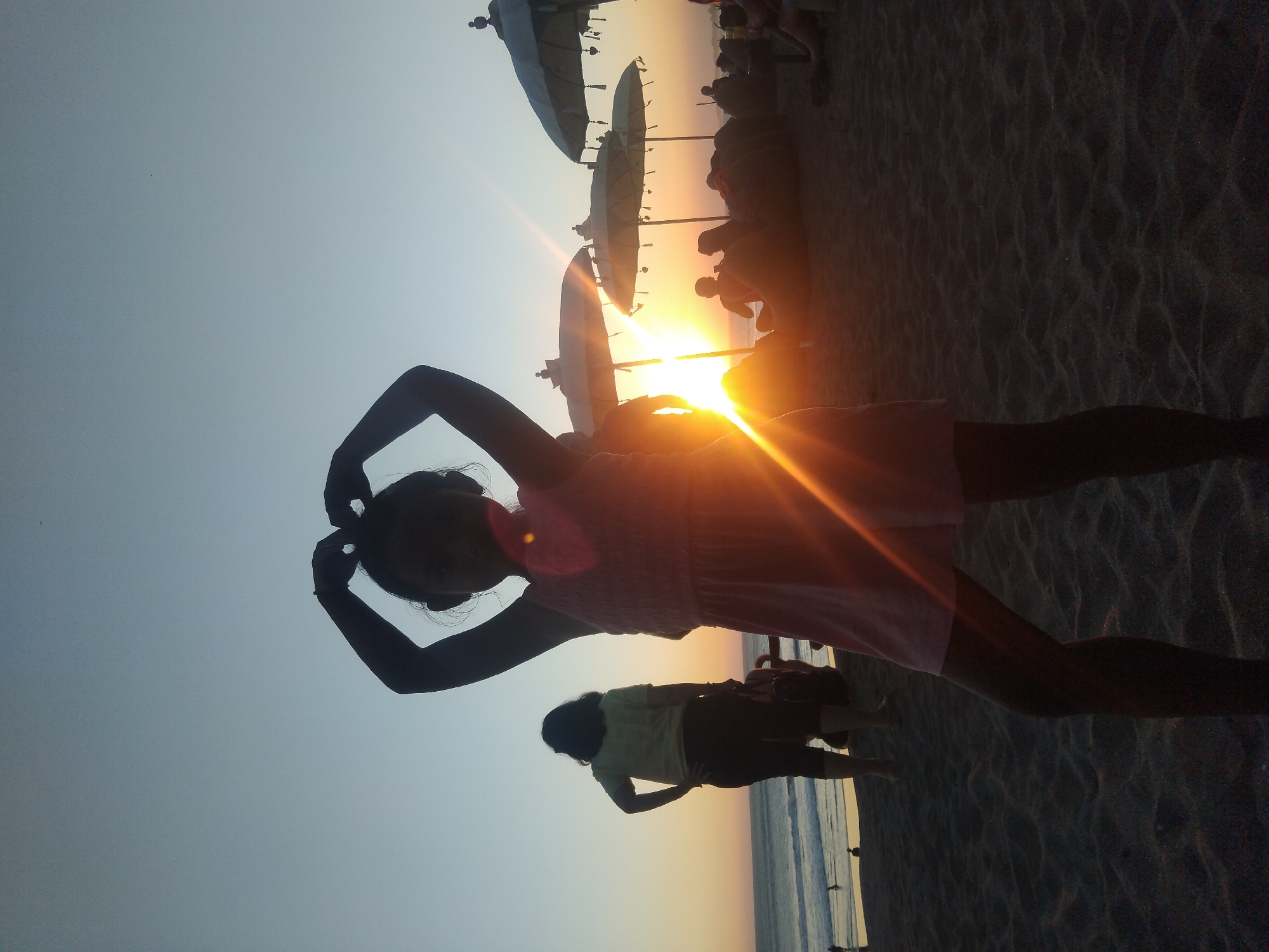 This vertical image (when correctly oriented) depicts a picturesque beach scene at sunset in North Kuta, Indonesia. The bright, setting sun is a central element, positioned just above the horizon on the middle-right, illuminating the sky with vibrant orange and yellow hues and creating dramatic silhouettes of the subjects.
In the foreground, a young person, likely female, is prominently silhouetted. She is wearing a light-colored dress and has her arms raised above her head, possibly adjusting her hair or posing. Her profile is visible against the brilliant sun, which also creates noticeable lens flares. Further back and slightly to her left, another individual is also silhouetted, standing on the sand and facing towards the ocean, appearing to wear a light top and darker bottoms.
The sandy beach occupies the lower portion of the frame, displaying subtle textures. In the mid-ground, a row of large, light-colored beach umbrellas is silhouetted against the vivid sunset, indicating a popular beach destination. Beneath and around these umbrellas, several other indistinct figures are visible, some seated, suggesting people are relaxing and enjoying the evening. The ocean's waterline is visible as a thin strip along the horizon, reflecting the ambient light.
The sky above transitions from the intense golden glow around the sun to a softer blue-grey further up. The strong backlighting and lens flares emphasize the silhouettes and the serene atmosphere of the sunset. There is no visible text in the image. The scene captures the tranquil beauty of a beach evening.