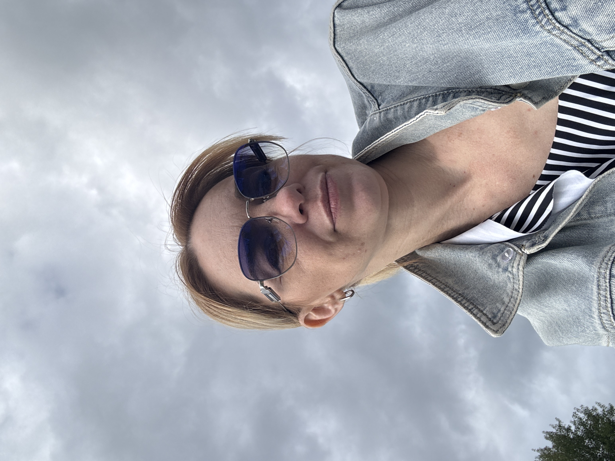 A low-angle, close-up, outdoor, waist-up photograph captures a Caucasian woman in Shuiskaya Chupa, Russia. She has short, blonde hair, fair skin, and is wearing large, silver-rimmed sunglasses with blue lenses. She has on a light blue denim jacket over a white shirt with a black and white striped pattern visible at the neckline. A silver hoop earring is worn in her visible left ear. The background consists of a cloudy, gray sky. The image quality is good.