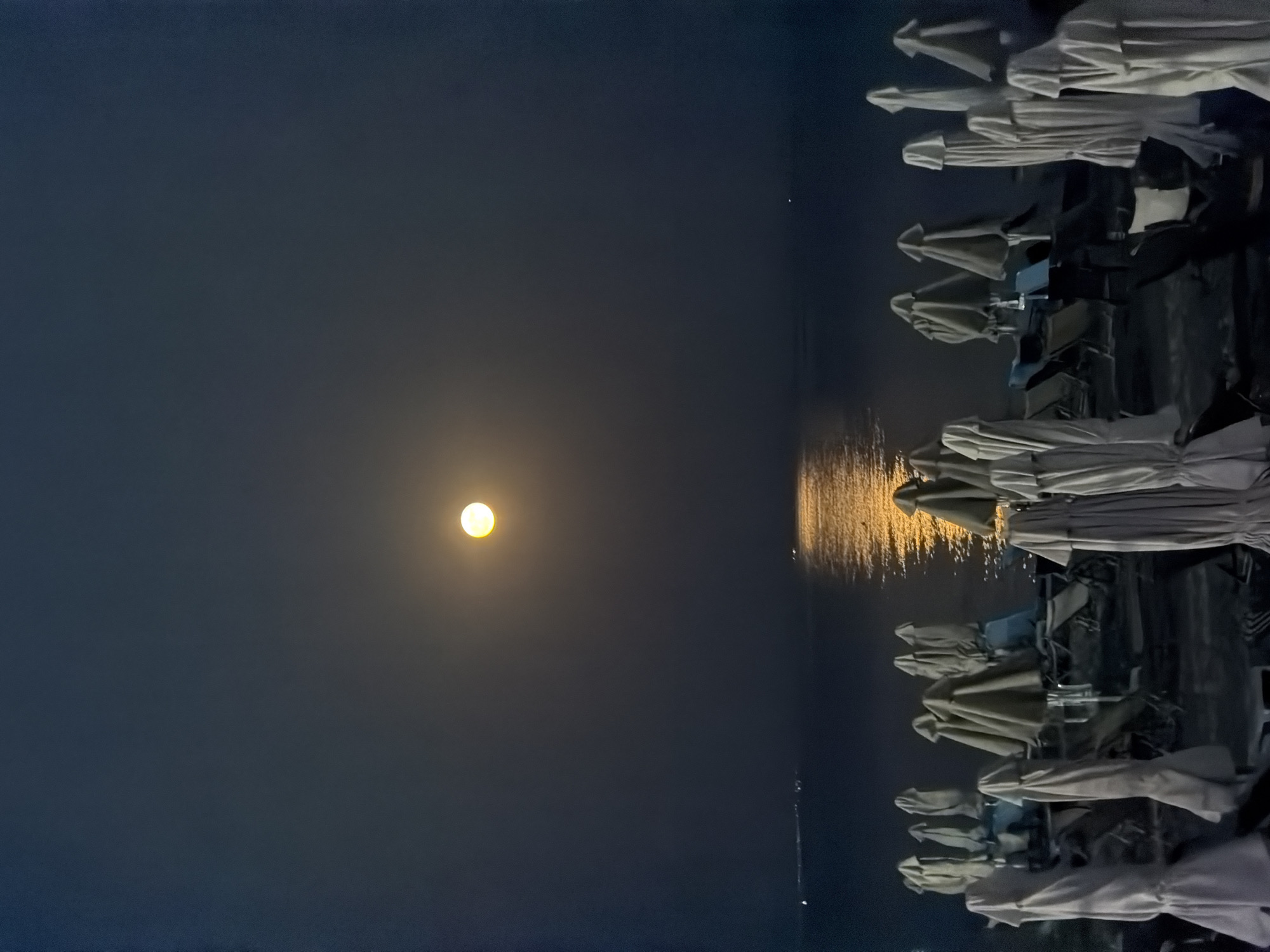 The image depicts a beach at night in Νέα Καλλικράτεια, Greece, under a full moon. The moon is prominent in the dark sky, casting a luminous reflection on the calm sea. In the foreground, numerous beach umbrellas are closed and covered, lined up with lounge chairs visible beneath them. The overall atmosphere is serene and tranquil.