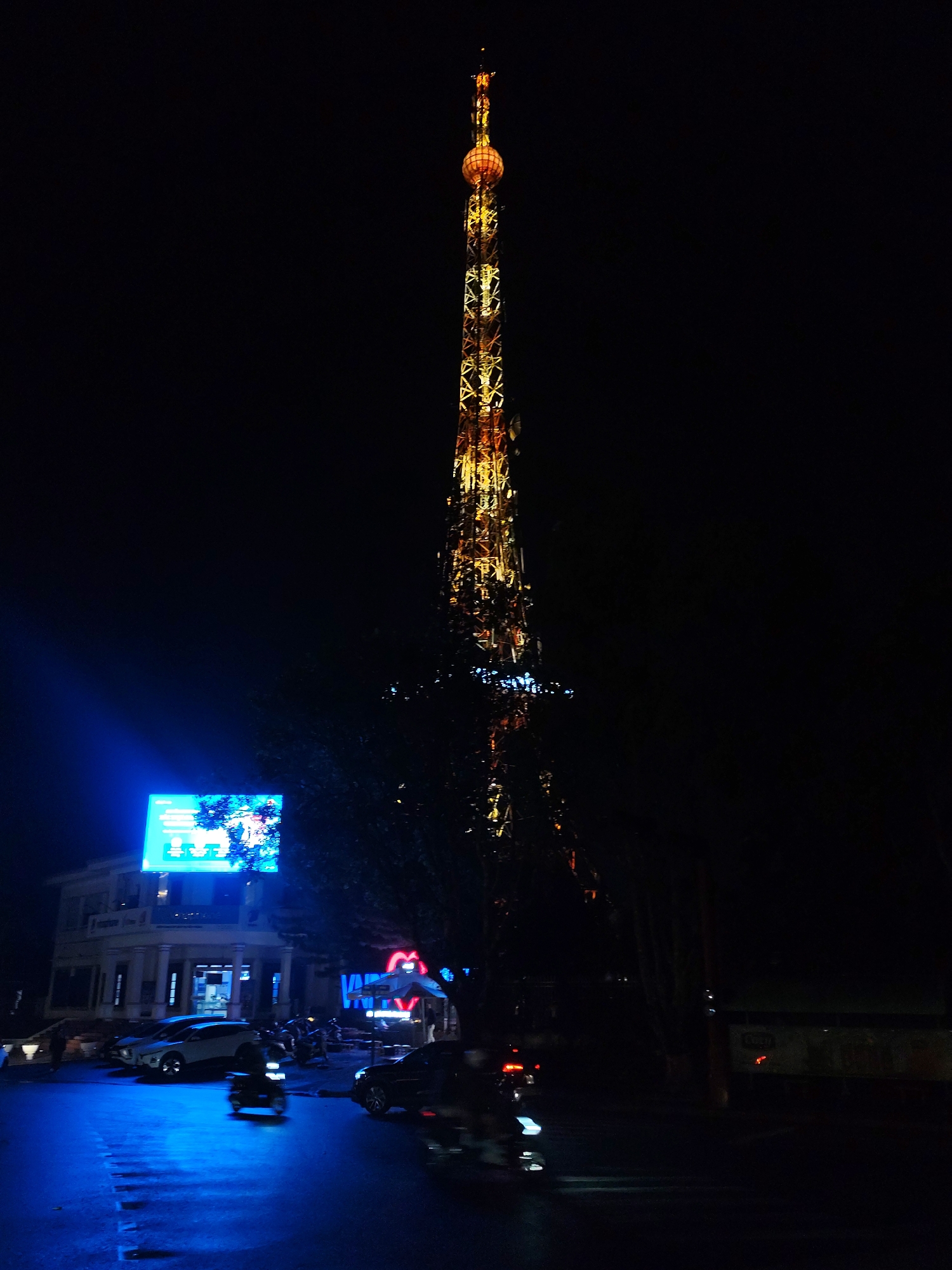 The image shows Da Lat TV Tower at night. The tower is tall, with a spherical structure at the top. It is illuminated with warm, yellow light. The lower portion of the tower is partially obscured by trees. In the foreground, a street scene is visible with moving vehicles, including motorbikes. The street is lit with blue light from a building facade and a brightly lit billboard, while other shops have neon signs. The overall environment is dark and night-time.