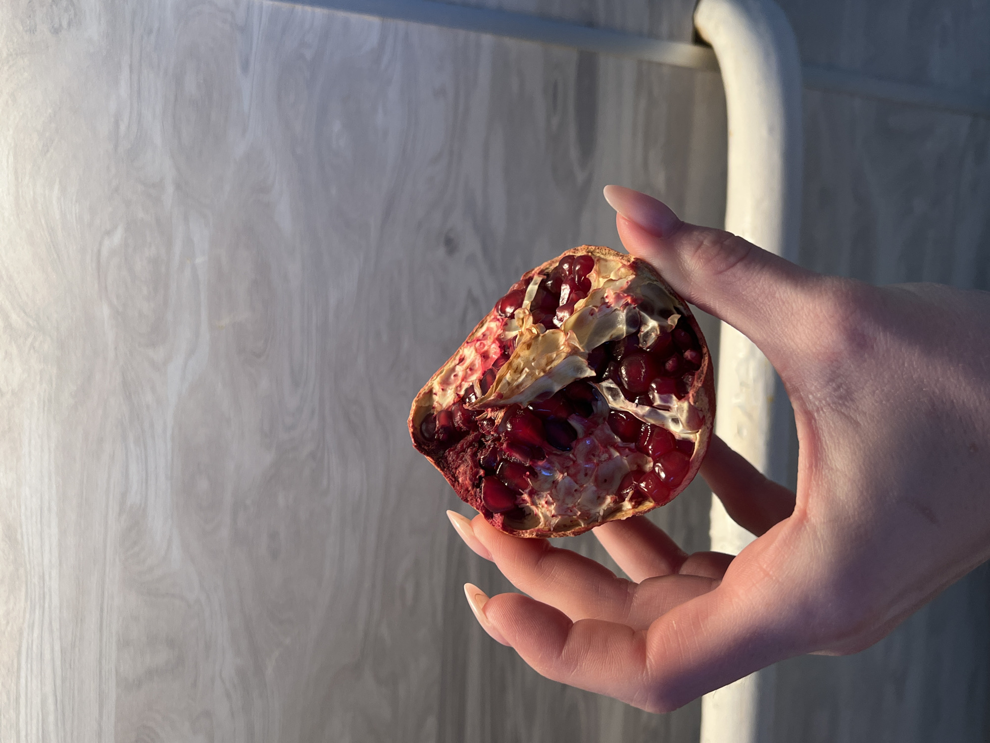 A human hand, likely female, holds a section of a red pomegranate against a light gray-brown background featuring a wood-grain pattern. The hand displays long, almond-shaped, light-colored fingernails. The pomegranate segment is split open, revealing dark red arils and white internal pith. A white, curved fixture or rail is partially visible on the right side of the frame. This scene is situated in the city of Chaikovsky, Russia.