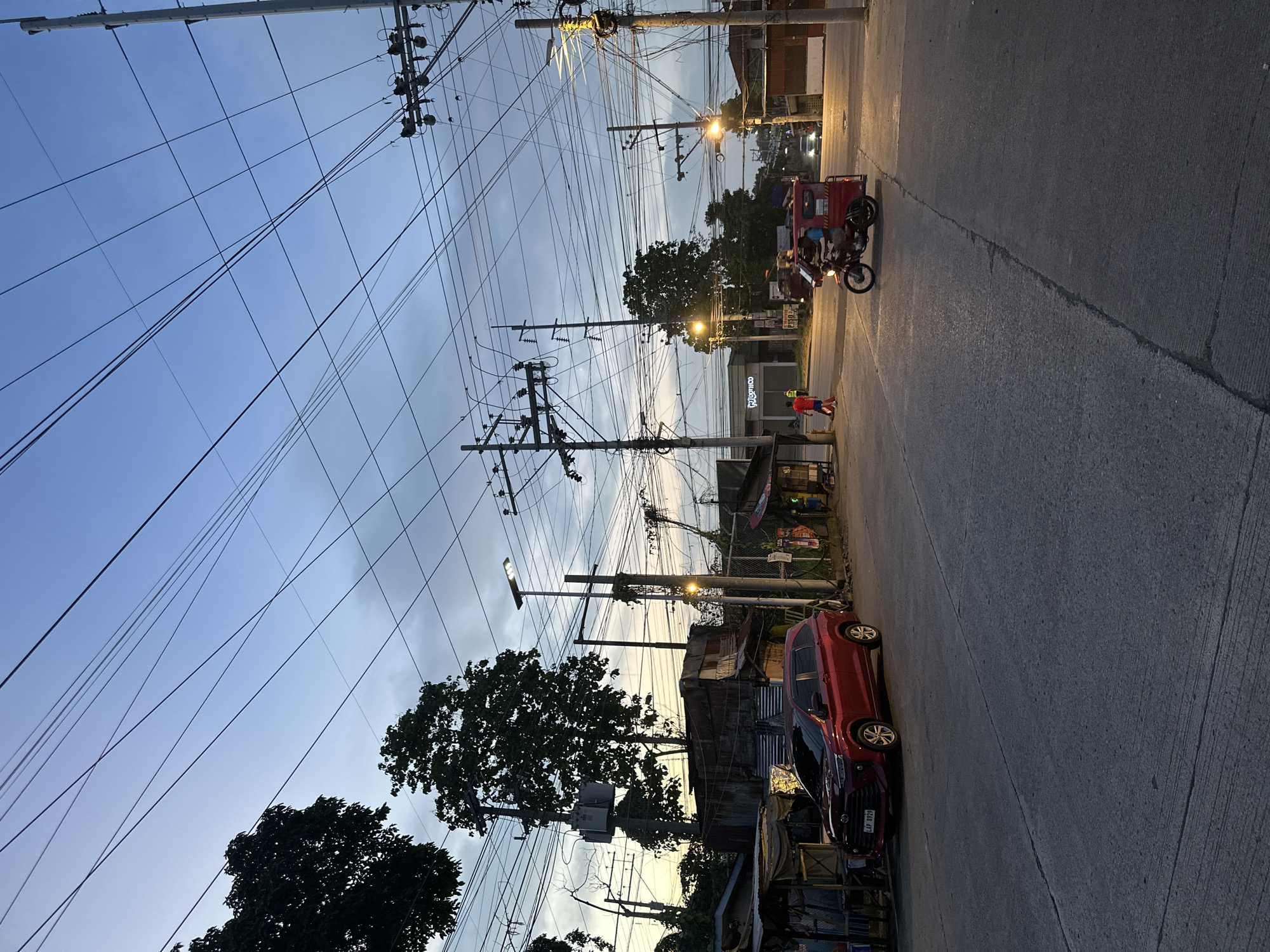 This image depicts a street scene in Davao City, Philippines, at dusk. The sky is a gradient of blue and gray, with scattered clouds, and is crisscrossed by a dense network of power lines supported by numerous utility poles. On the left side of the road, a red SUV is parked in front of structures that appear to be small shops or dwellings with corrugated metal roofing. A few people are visible on the sidewalk and street. To the right, a motorized tricycle with a passenger is traveling on the road, illuminated by streetlights. The overall impression is of a typical urban environment during the transition from day to night, characterized by overhead infrastructure and active street life.