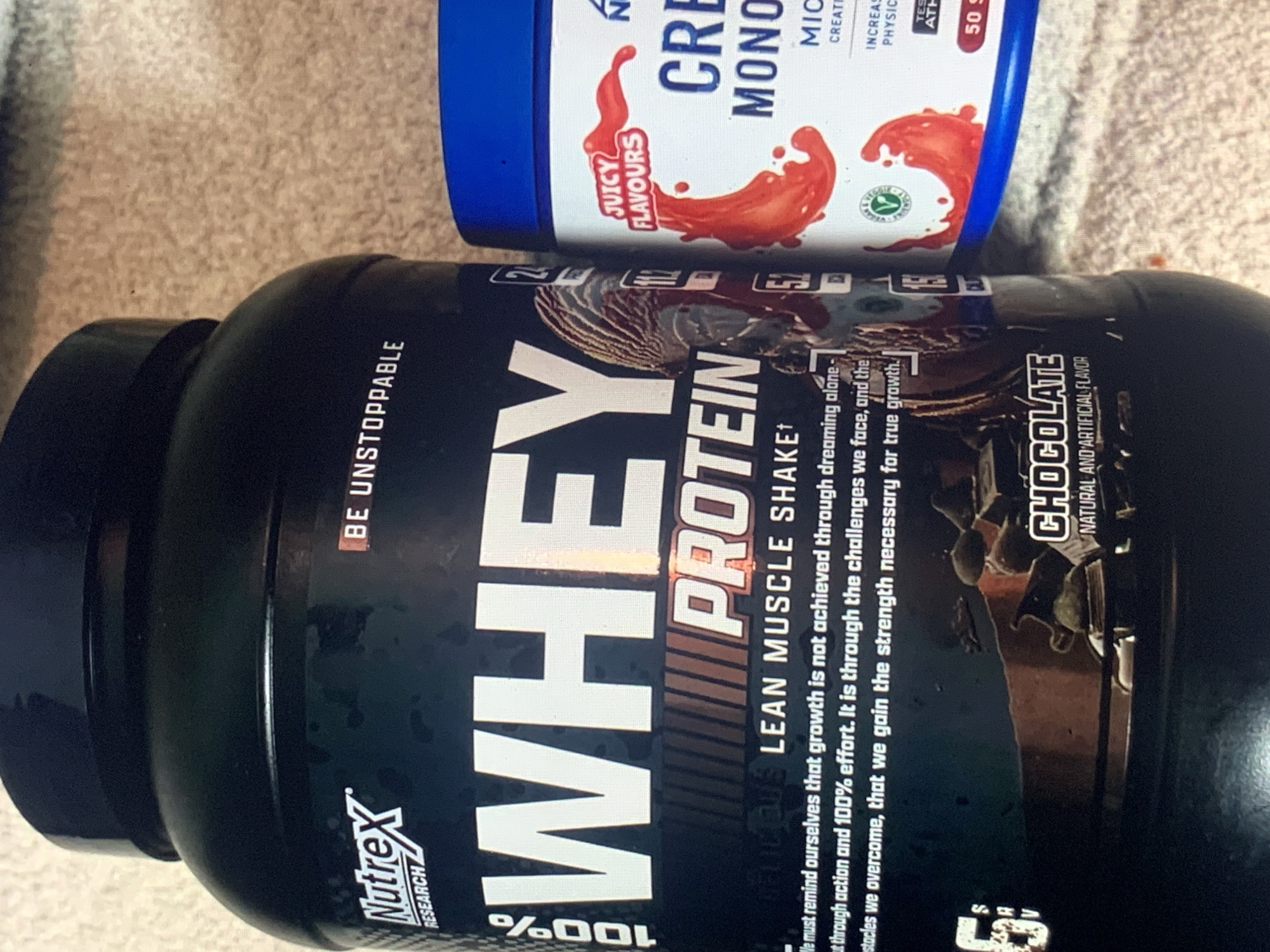 An image captures two dietary supplement containers resting on a textured, light brown surface, possibly a carpet or rug, with the objects oriented sideways.
The prominent item is a large, black tub of "WHEY PROTEIN" by "Nutrex RESEARCH". The flavor is "CHOCOLATE NATURAL AND ARTIFICIAL FLAVOR," and it is described as a "LEAN MUSCLE SHAKE." A motivational slogan reads, "BE UNSTOPPABLE," and additional text states, "We must remind ourselves that growth is not achieved through dreaming alone - but through action and 100% effort. It is through the challenges we face, and the obstacles we overcome, that we gain the strength necessary for true growth." The label also features "100%" and an illustration of a bodybuilder.
Behind and slightly to the right of the whey protein tub is a smaller, blue-lidded, white tub. This product is "CREATINE MONO," likely "Micronized Creatine Monohydrate." The label highlights "JUICY FLAVOURS" with a red splash graphic, and mentions "INCREASES PHYSICAL PERFORMANCE" and "TESTED ATHLETE." It indicates "50 SERVINGS" and has a small green circular logo. The brand initial "N" is visible, suggesting it's also a Nutrex product.
The scene is indoors, presenting a clear view of the product details. No activity is taking place. The overall lighting is even.