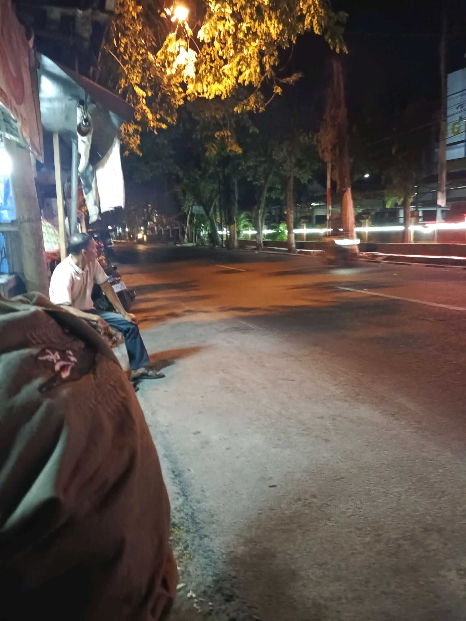 An adult male, wearing a light-colored collared shirt and dark pants, is seated on a motorcycle parked on the side of a paved road at night. He is looking to his left. The road is illuminated by streetlights and the headlights of passing vehicles, which appear as streaks of light due to motion blur. Trees line the side of the road, and some structures, possibly shops or stalls, are visible in the background. The scene takes place in Lamongan, Indonesia.