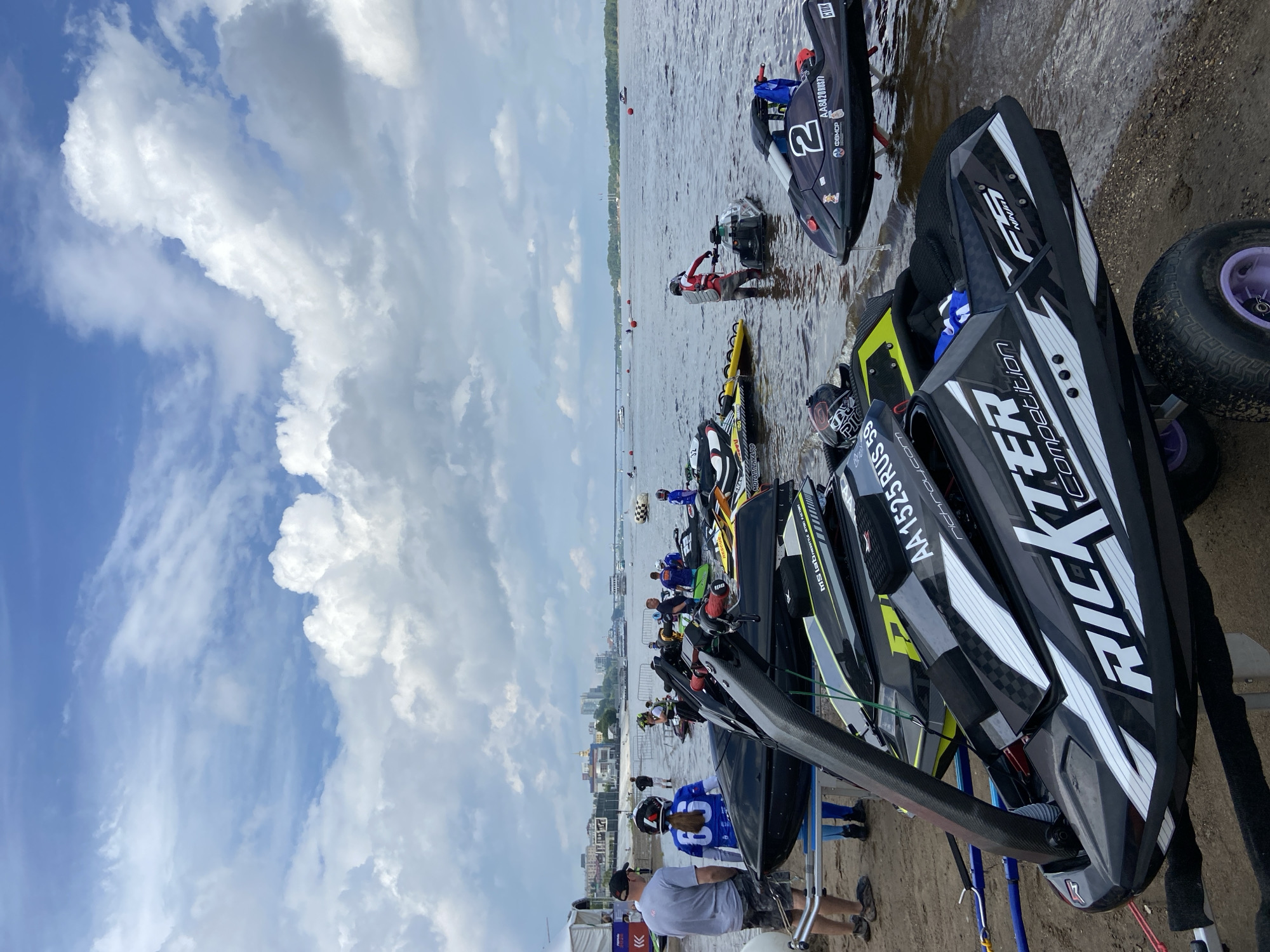 A sunny day with scattered clouds over a wide river, likely the Kama River in Perm, Russia, where a jet ski competition is about to begin. In the foreground, several jet skis are lined up on the sandy shore. The closest jet ski, a black and white "RICKTER Competition" model, is on a trailer with large tires. Further back, other jet skis, including one with the number "2" and "AA8420RUS77" painted on its hull, are partially submerged in the shallow water. Several people are present, some in competition gear like helmets and life vests, while others are dressed casually. A person in a red and grey suit is standing by a jet ski in the water. In the background, a city skyline with buildings and a bridge can be seen across the river. The overall atmosphere suggests a sporting event in progress.