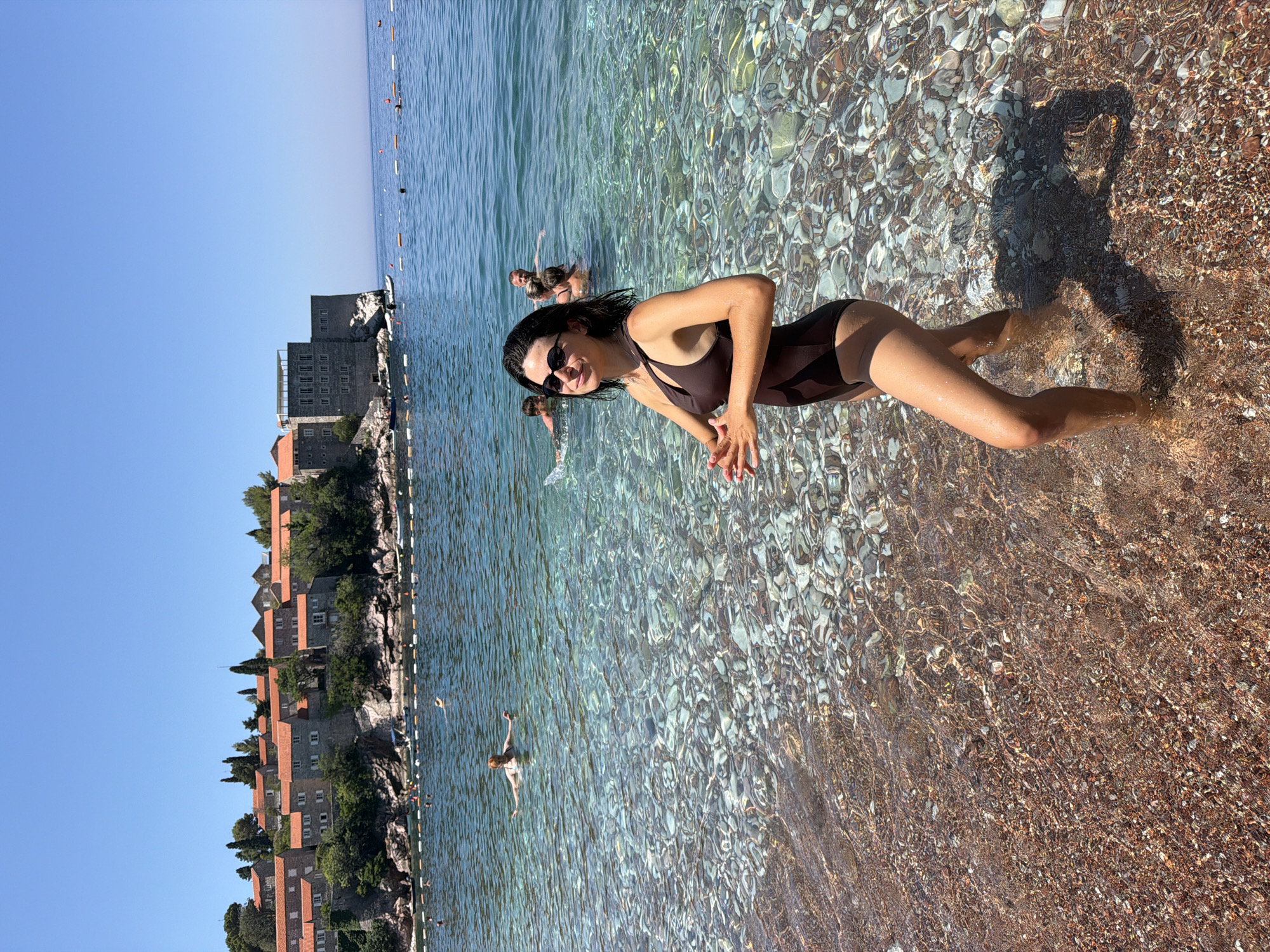 A woman wearing a brown one-piece swimsuit stands in shallow, clear water on a pebble beach. She has dark hair, sunglasses, and is looking towards the camera with her hands clasped in front of her. In the background, other people are swimming and wading in the sea. Behind them, a stone village with red-tiled roofs is situated on a cliff overlooking the water. The sky is clear and blue. The location is Sveti Stefan, near Rađenovići, Montenegro.