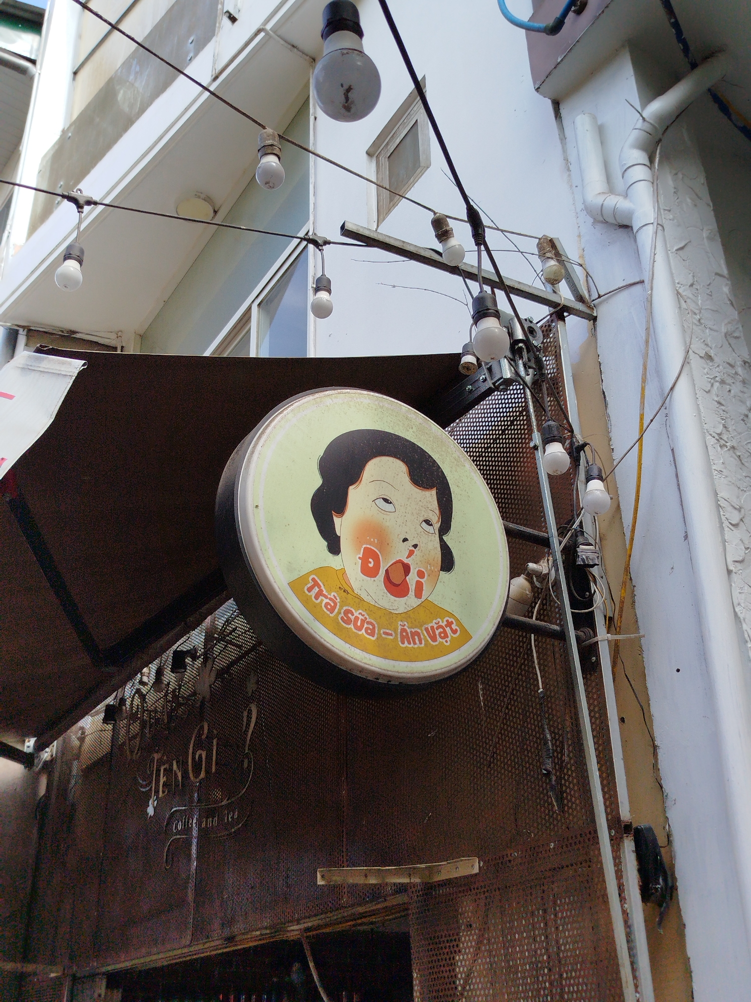 The image shows the exterior of a café named "Ten Gi?" in Da Lat, Vietnam. The café is adorned with a retro sign featuring a cartoon face with a large, open mouth, and Vietnamese text below: "Trà sữa - Ăn vặt" (Milk tea - Snacks). The café's name is displayed on a perforated metal sheet in a decorative script. A string of bare lightbulbs hangs above the entrance. The buildings surrounding the cafe are white and somewhat weathered. A drain pipe runs along the side of the building to the right of the cafe entrance.