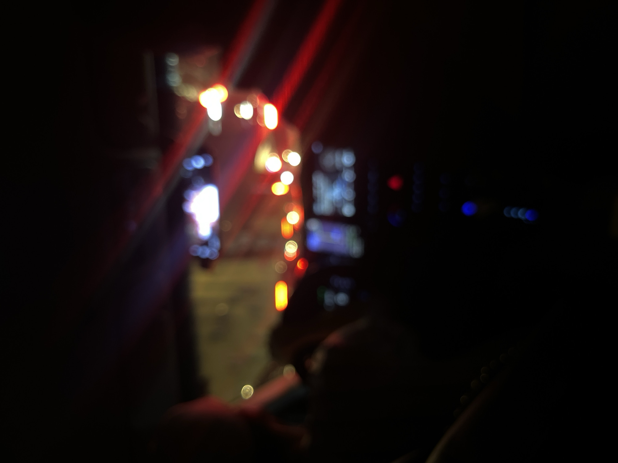 The image appears to be taken from inside a car at night, possibly in Moscow, Russia given the presence of Cyrillic script on a screen within the vehicle. The focus is soft, creating bokeh effects with the lights. Outside the car, there are strings of red and white lights, likely traffic lights or building illuminations. Inside the vehicle, a dashboard with a display screen is visible. Other details are obscured by darkness.