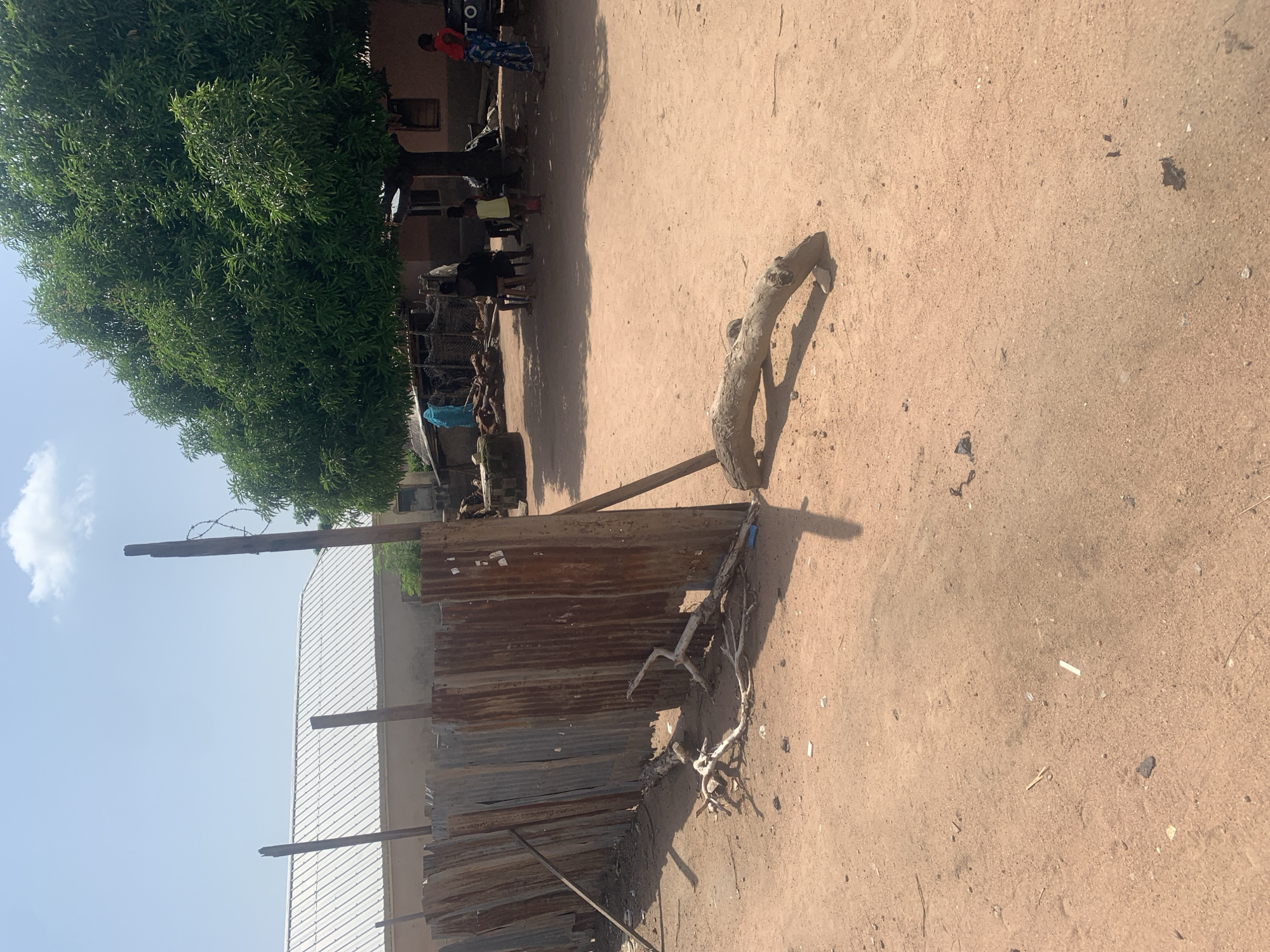 The image depicts an outdoor scene in Jalingo, Nigeria, characterized by a dusty, reddish-brown ground and clear blue skies with scattered clouds. On the left, a fence constructed from corrugated metal sheets, showing signs of rust and age, runs across the frame. Several wooden poles, some with barbed wire, support the fence. A large, leafy tree dominates the right side of the image, providing shade to the area beneath it.
Several people are visible in the background. One person in a bright blue garment stands near a structure. Under the tree, a group of individuals are gathered, some sitting on stools. A small child in a yellow dress is present, and another child is standing next to them. A person in a red top and blue patterned skirt is also visible. To the far right, the rear of a vehicle with the partial word "TOY" visible on it is parked. Various objects, including pieces of wood and debris, are scattered on the ground. The overall environment suggests a rural or semi-urban setting in Jalingo.