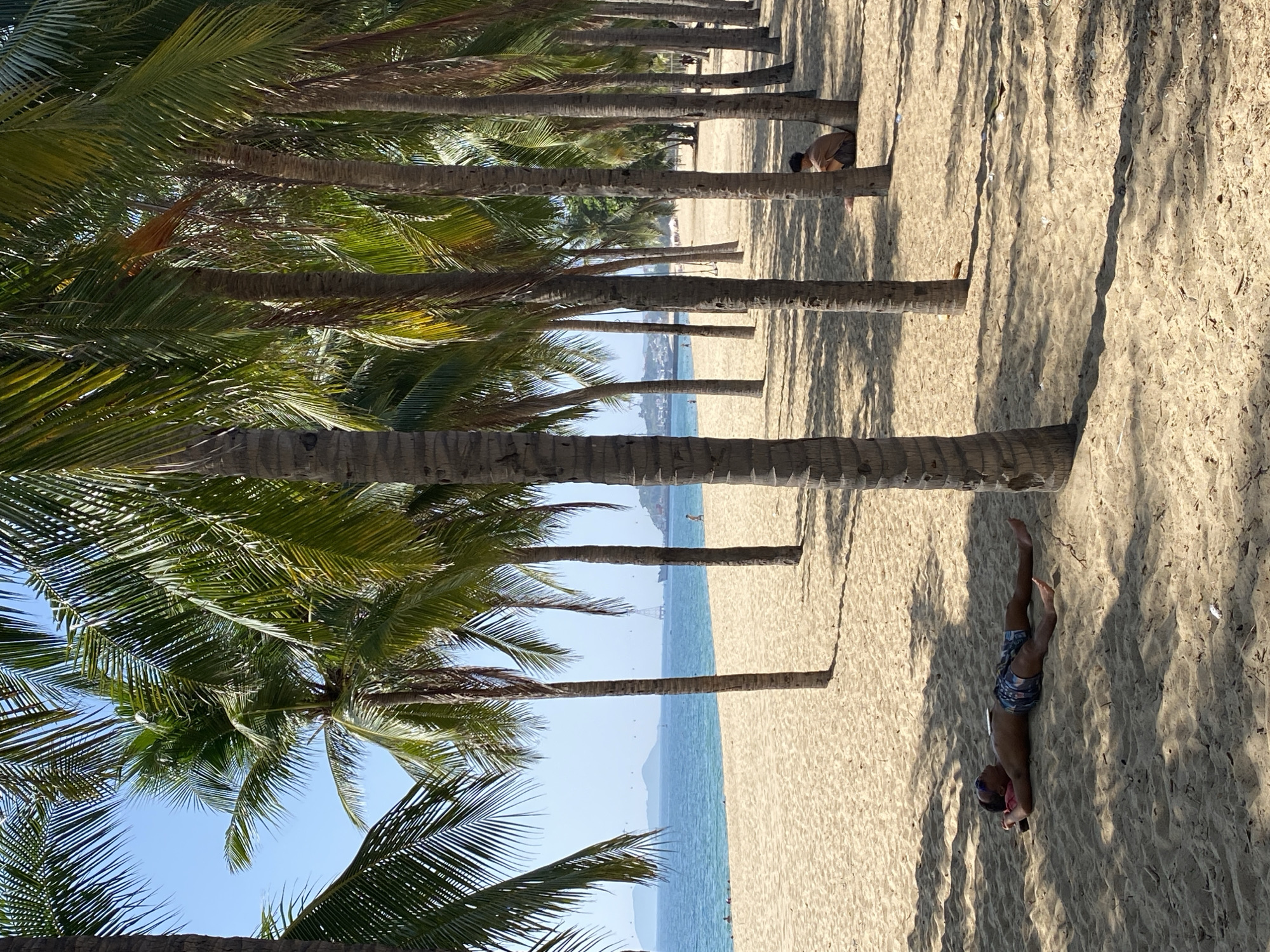 A man lies on his back on a sandy beach in Nha Trang, Vietnam, with his head on a red object and wearing patterned shorts. He appears to be sunbathing or resting under the shade of palm trees that line the beach. In the distance, the ocean is visible, with a few boats and islands on the horizon. Another person sits under a palm tree further down the beach. The sand is light brown and appears smooth, with shadows cast by the palm trees creating a dappled effect. The sky is clear and blue.