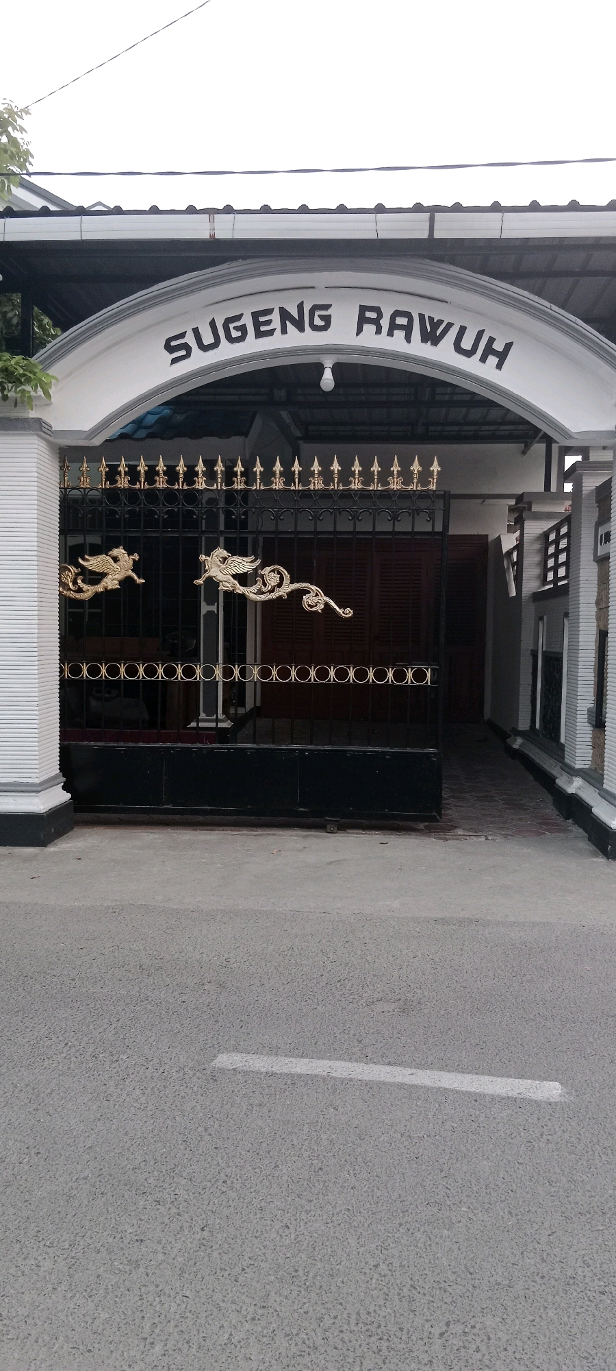 The image shows the entrance to a property with an arched gate. The archway has the text "SUGENG RAWUH" inscribed on it in dark, block letters. The gate itself is made of black metal bars with decorative golden accents, including what appear to be two stylized griffins or winged creatures. The gate is closed, obscuring the view of the property beyond, though a red-brown wooden door is partially visible through the bars. To the left of the gate, there is a white pillar with a textured finish. The ground in front of the gate is paved with asphalt. The environment appears to be outdoors, with a building structure visible behind the gate and a bit of greenery on the left side. The lighting suggests it is daytime, possibly overcast. The location is identified as Soko, Indonesia.