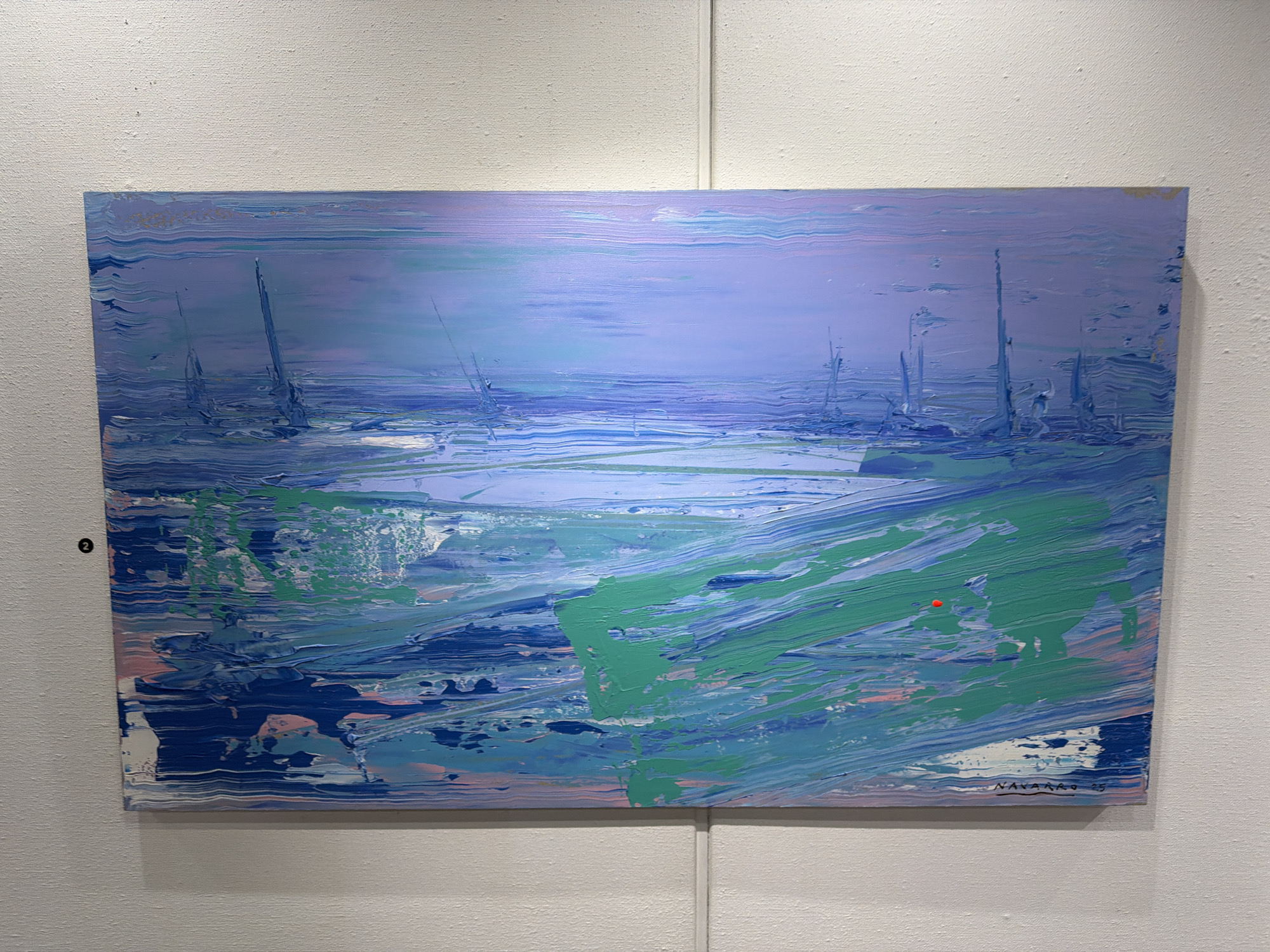 The image displays an abstract oil painting on canvas, depicting a seascape with a lavender sky and a turbulent sea. The painting is characterized by thick, gestural brushstrokes in shades of blue, green, pink, and white, creating a sense of movement and texture. Several vertical elements, possibly representing sailboats or masts, are visible in the distance. A small red dot is present in the upper right quadrant of the painting, and another is on the canvas near the bottom right. The artist's signature, "NAVARRO '25," is visible in the lower right corner. The painting is hung on a textured, off-white wall. To the left of the painting, a small circular sticker with the number "2" is attached to the wall. This artwork is displayed at the Looping Gallery, located on Rua das Flores 7, in the historic center of Cascais, Portugal, a town known for its coastal charm and artistic heritage.
