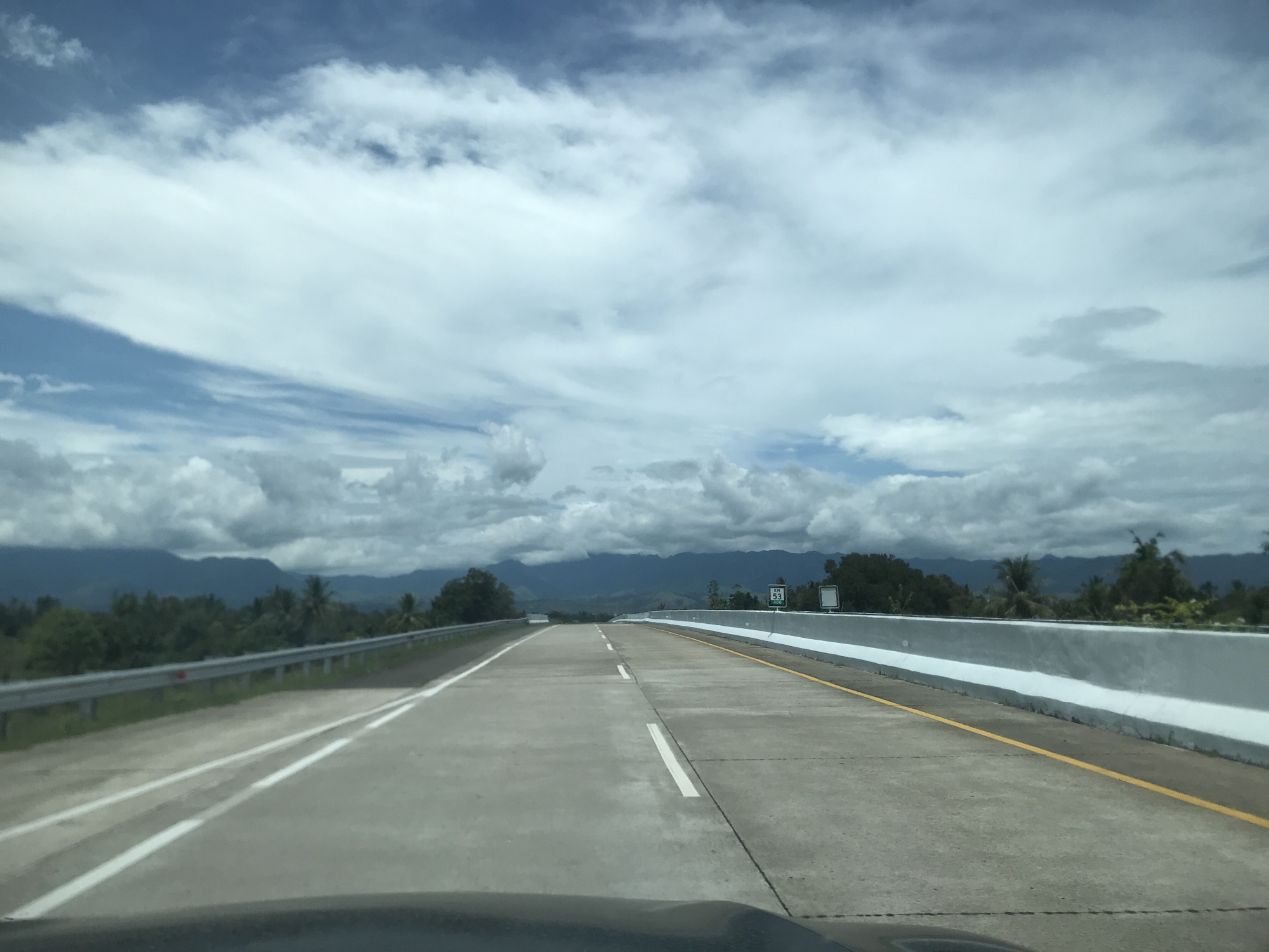 The image depicts a view from a vehicle driving on a highway in Cotkareung, Indonesia. The road is a concrete multi-lane highway with a center divider and yellow lines marking the right side. A gray metal guardrail is on the left side, and a concrete barrier is on the right. A green sign with "KM 55" is visible on the right side of the road. In the background, there are green trees and hills under a partly cloudy sky. The sky is primarily blue with white clouds. Part of the dashboard of the vehicle is visible at the bottom of the image.