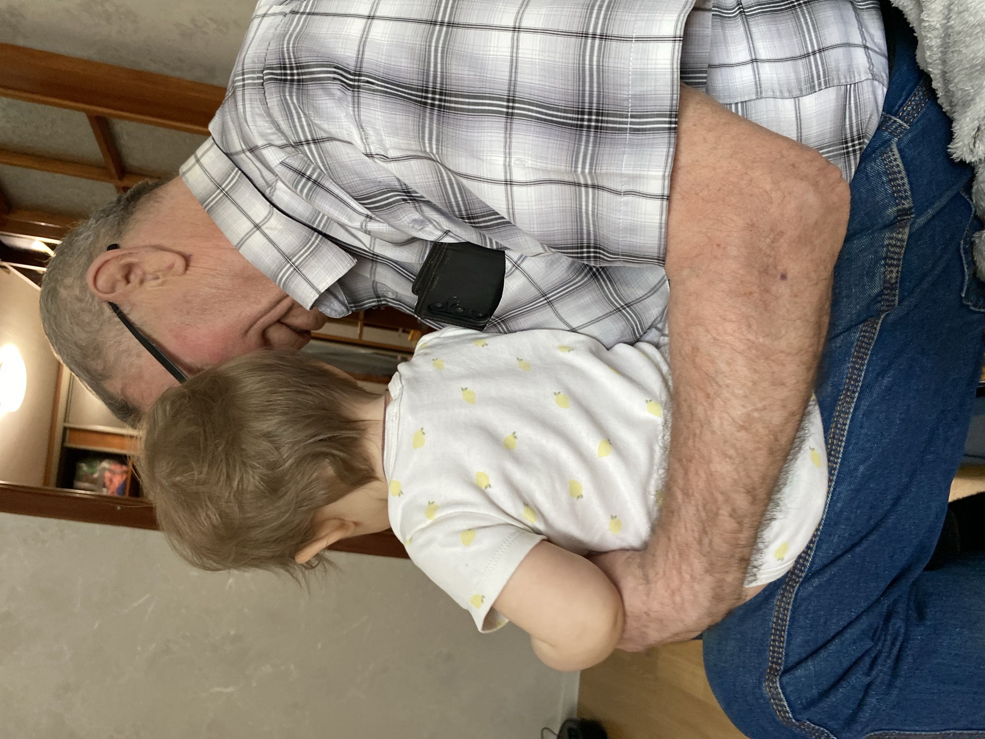 In a residential setting in Chaikovsky, Russia, an adult male holds an infant on his lap. The adult, visible from the mid-torso to the head, is an older male with short, grey hair and black-framed eyeglasses. He is dressed in a light-colored plaid button-up shirt with grey and white patterns, and dark blue jeans. A dark, possibly leather, object is partially visible in his left shirt pocket. His right arm is wrapped around the infant. The infant, seen from the back, has light brown hair and wears a white short-sleeved top adorned with multiple small yellow lemon patterns. Both individuals are looking forward and slightly downwards, away from the camera. The floor beneath them is light-colored wood. The background includes a light-colored, possibly textured wall to the left and a vertical wooden structure to the right, suggesting an indoor domestic environment.