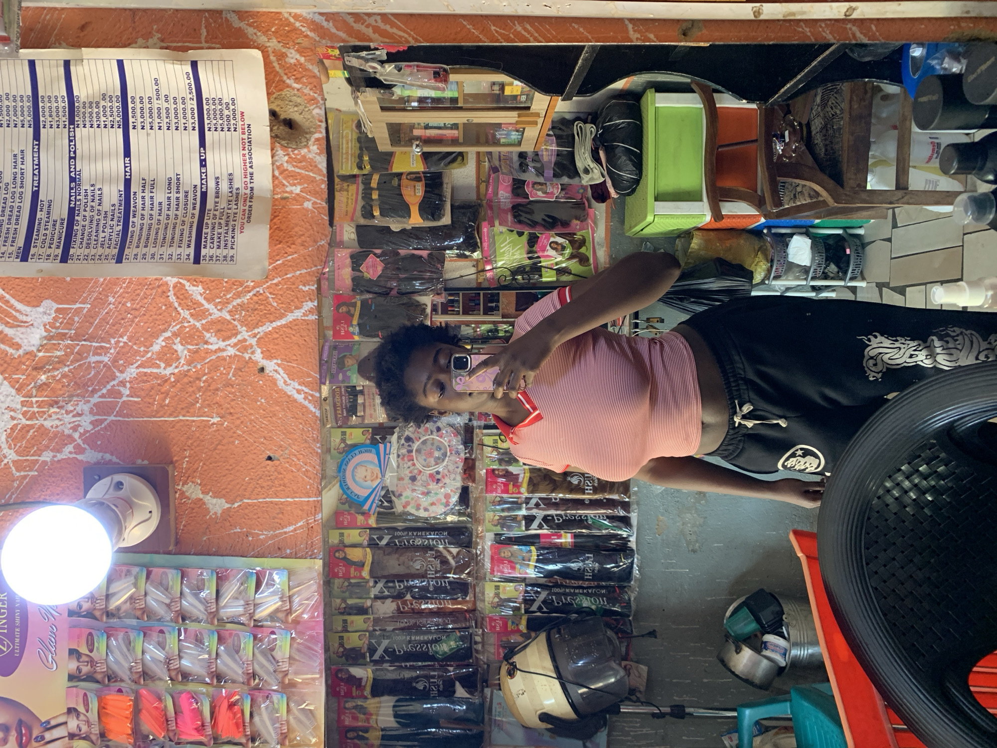 A young Black woman is taking a mirror selfie in what appears to be a hair and beauty salon. The woman is wearing a pink and white striped crop top and black sweatpants with a white graphic design. She is holding a pink smartphone. Behind her, the salon is filled with shelves of hair products, including wigs and extensions, as well as nail products and other beauty supplies. A price list for services is visible on the wall, listing treatments like "Steaming Hot," "Cold Steaming," "Manicure," and "Hair Weaving." The salon's walls are an orange, textured paint with white splatters. A white light bulb is illuminated on the ceiling. The overall scene suggests a small, local business in Rafin Par, Nigeria, offering a range of hair and beauty services.